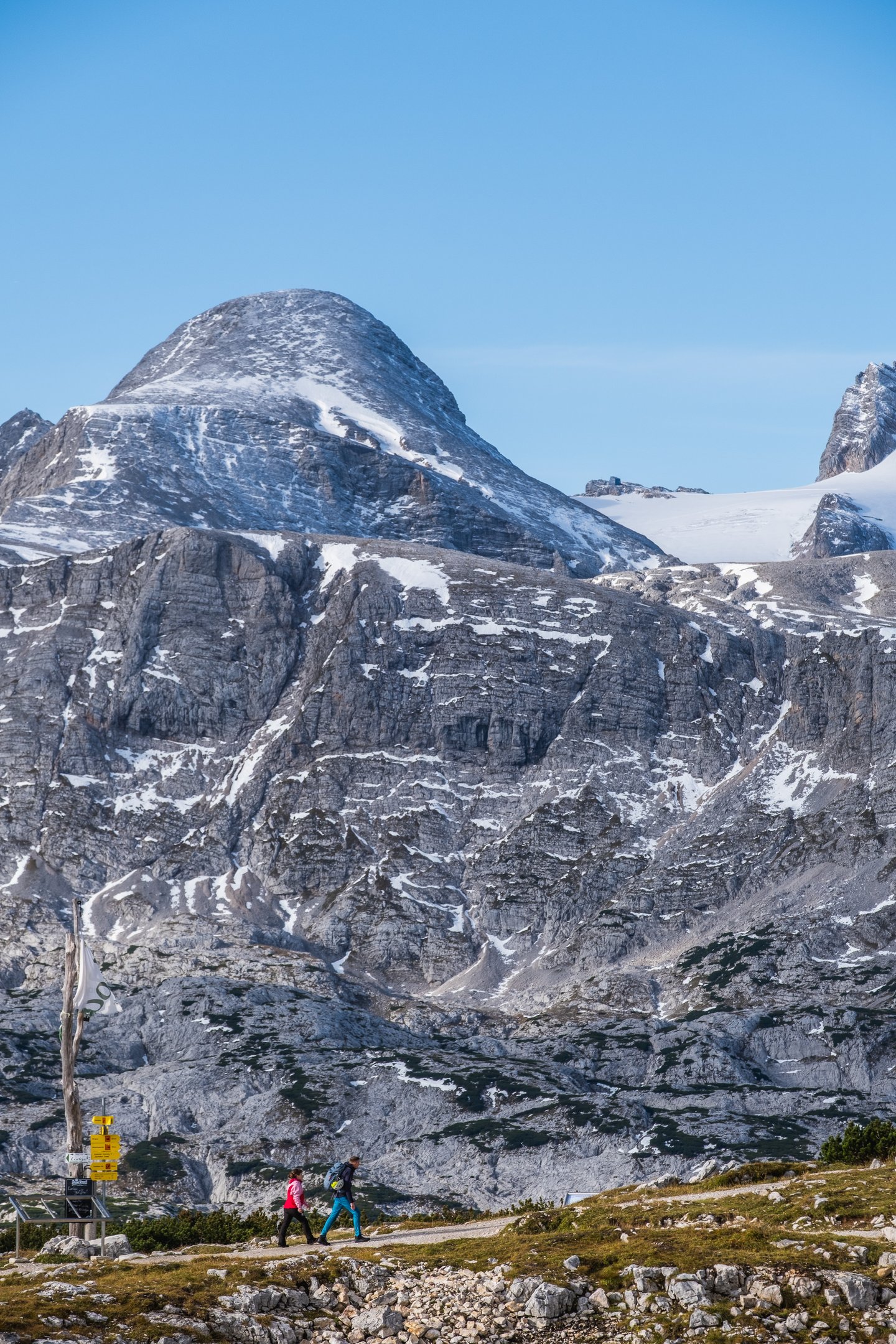 Two hikers walk a gravel plateau path, dwarfed by the grey limestone wall of the Dachstein massif with a snow-dusted rounded summit and a glacier edge above them.