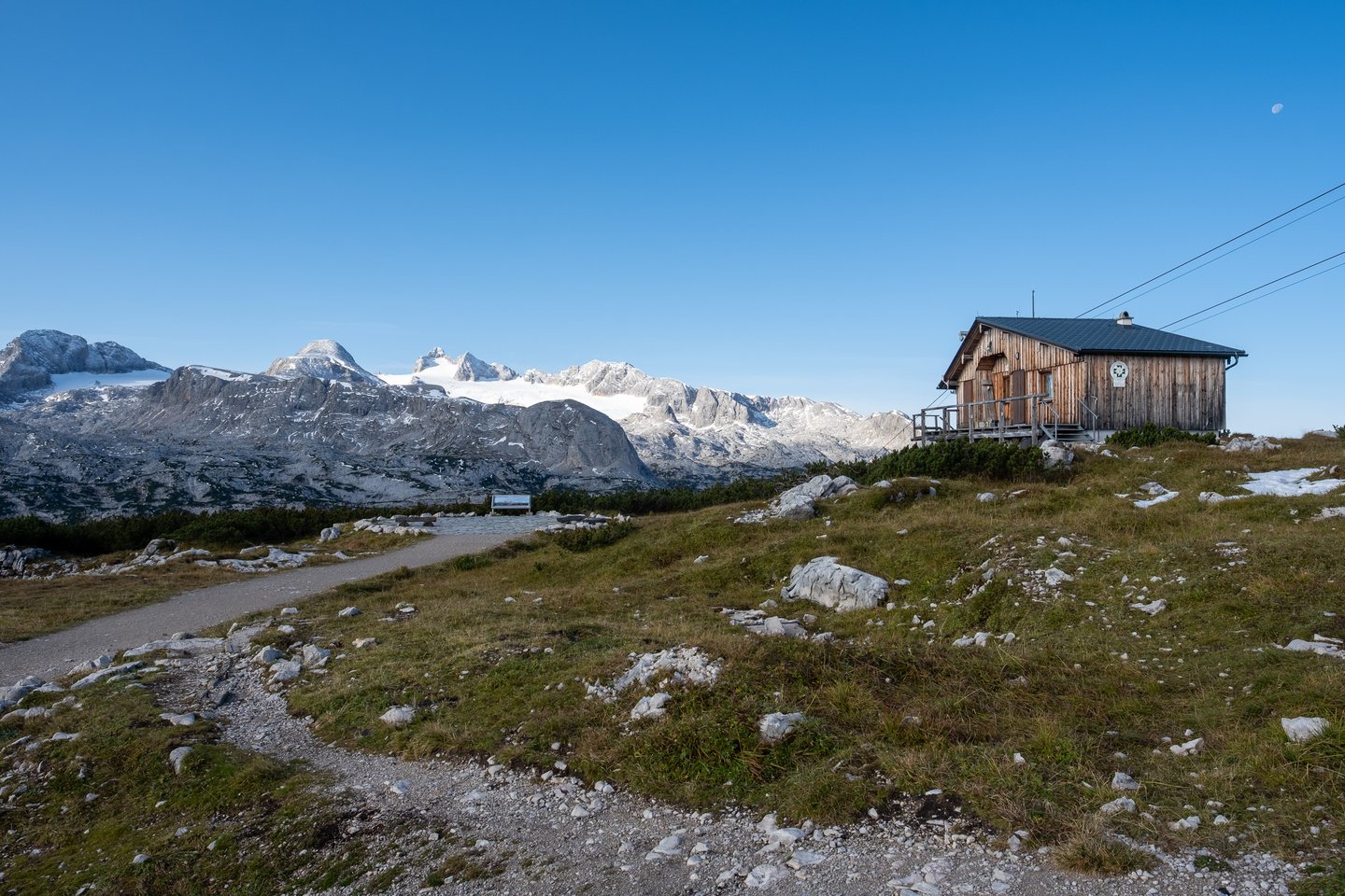 A small weathered wooden hut sits on the grassy Krippenstein plateau in early morning light, facing the long snow-topped wall of the Dachstein range with a pale moon overhead.