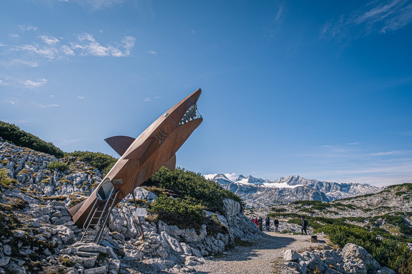 A rust-red steel sculpture shaped like a shark breaching out of the limestone rears above a gravel path on the Krippenstein, with the glaciated Dachstein summits behind it under a clear sky.