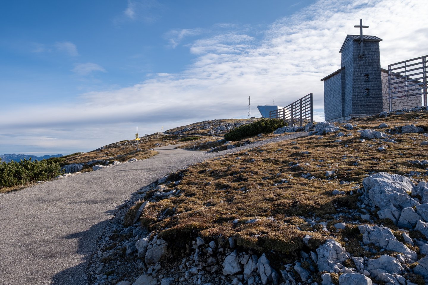 A shingled chapel tower topped by a cross stands on the Krippenstein ridge above a curving path, with the angular metal Welterbespirale viewing platform on the skyline behind it.