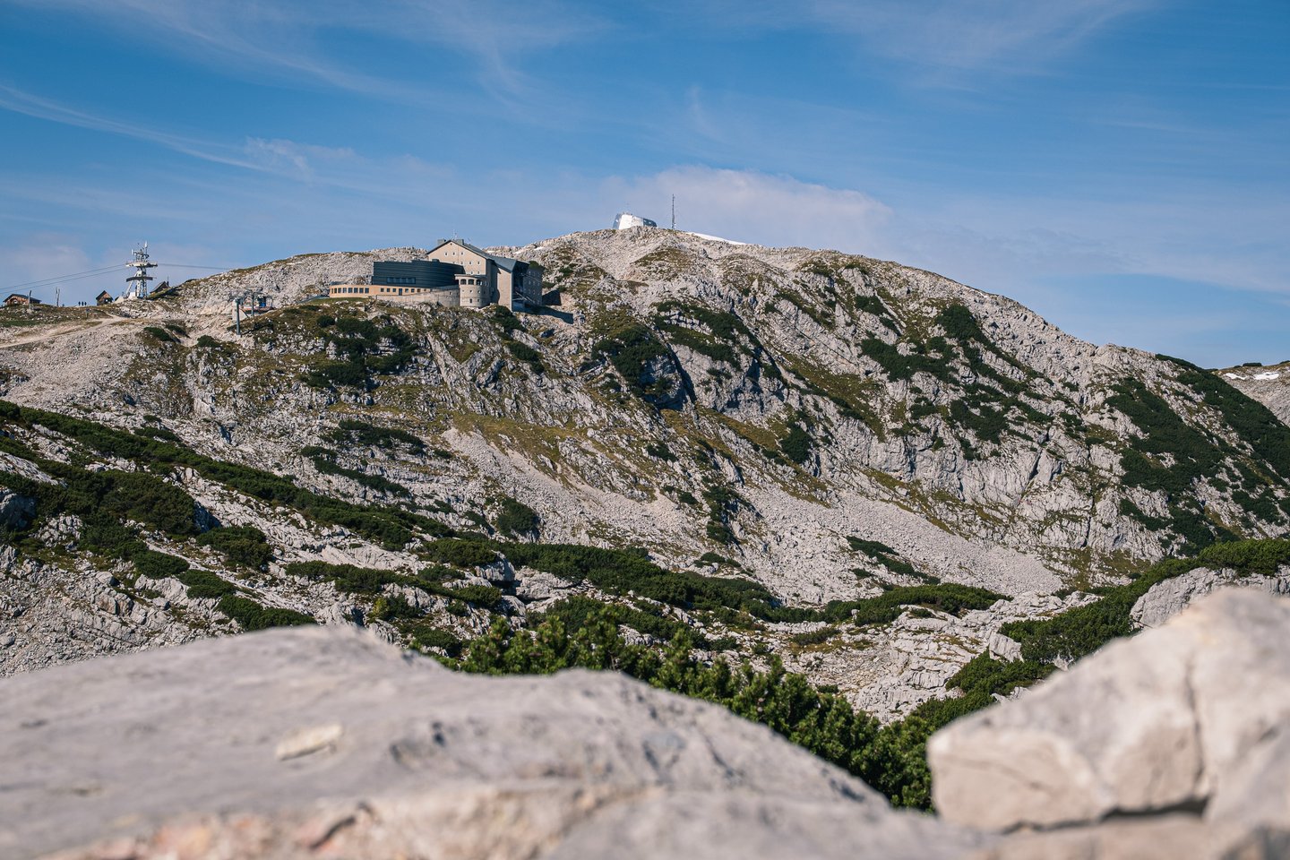 The Krippenstein summit ridge with its cable car top station and long lodge building, seen from across a bowl of pale karst rock and dwarf pine, with a blurred boulder in the foreground.