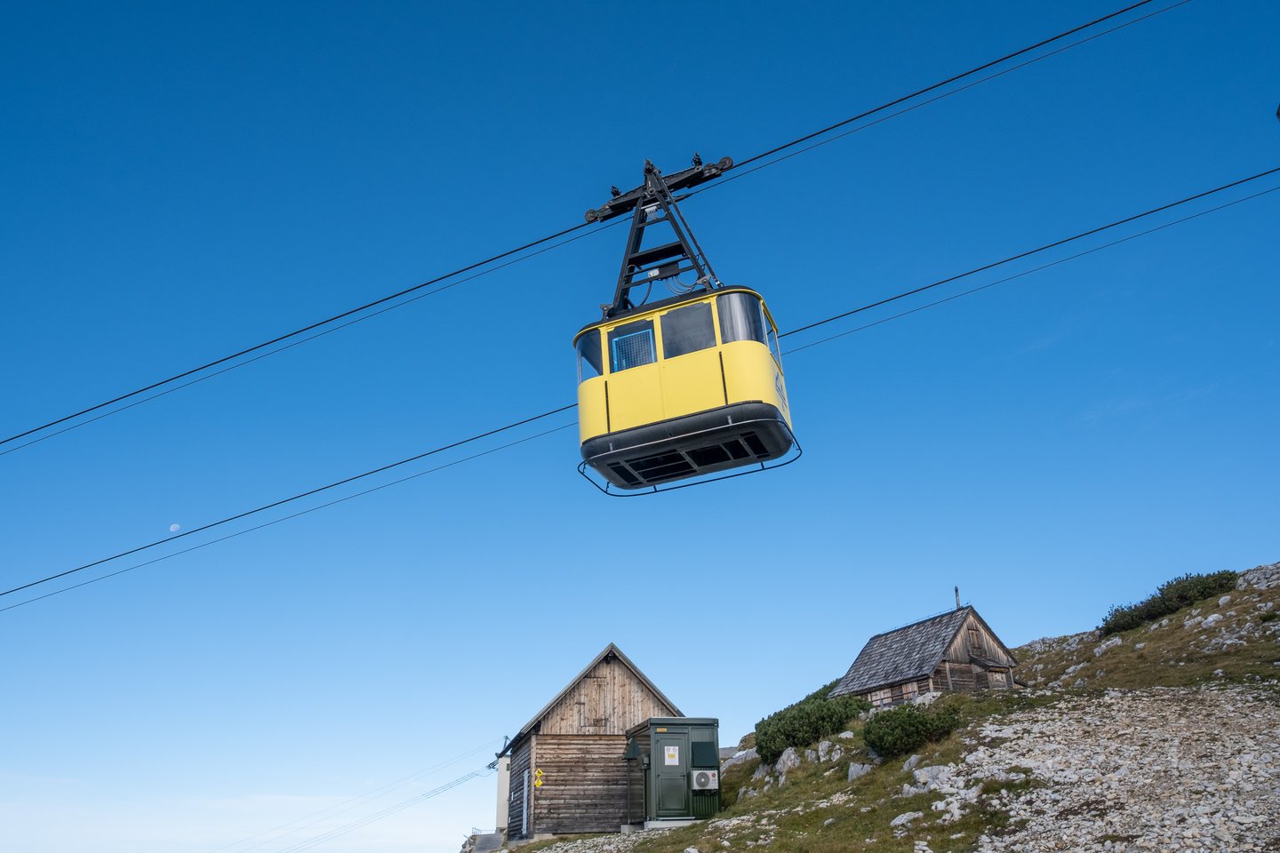 Looking straight up at a yellow Dachsteinbahn cabin gliding along its cables against a deep blue sky, with two wooden huts on the ridge below.