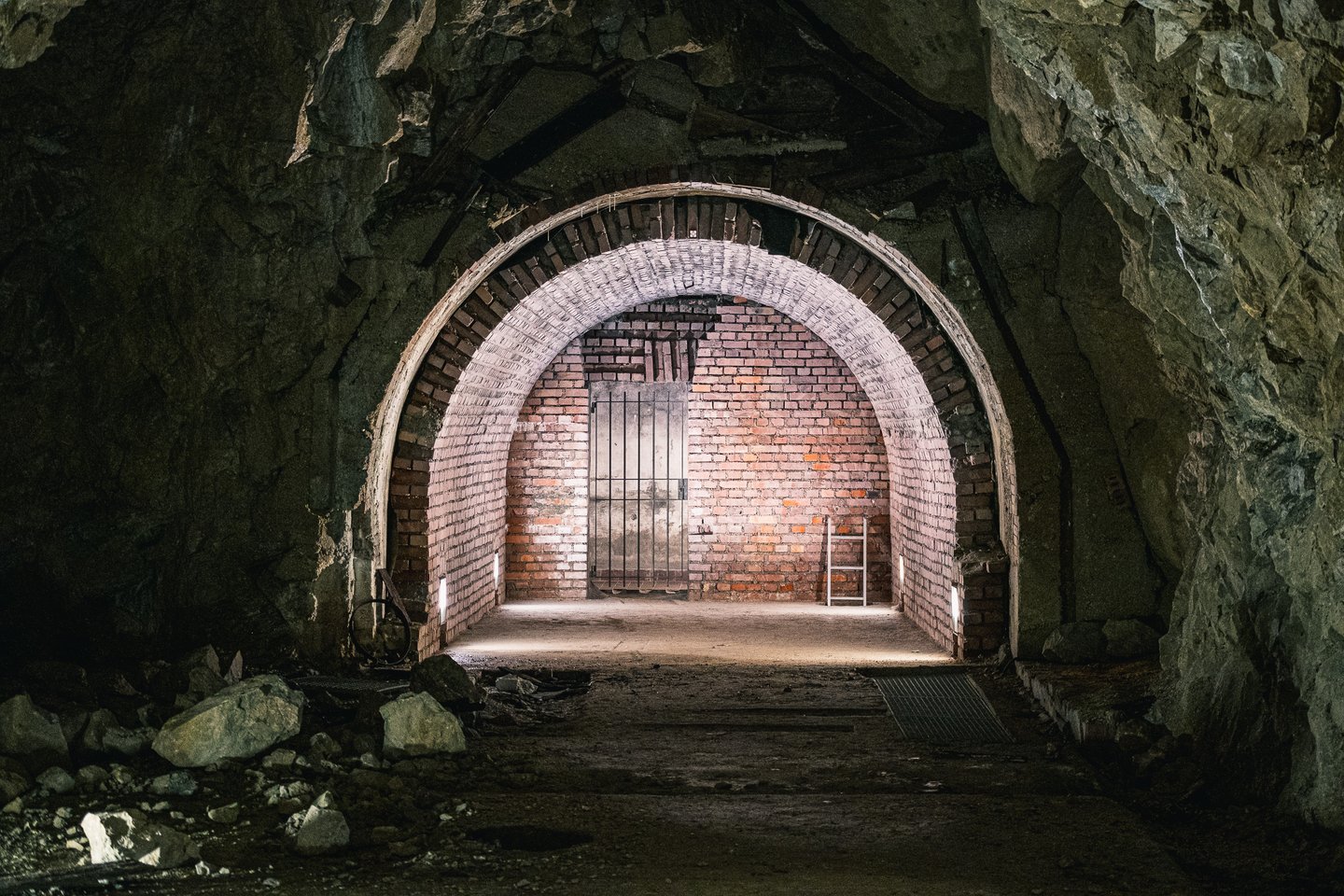 A brick lined arched portal set into raw blast rock in the Obersalzberg bunker system, lit from the floor and closed at the far end by a steel barred door, with rubble in the foreground.