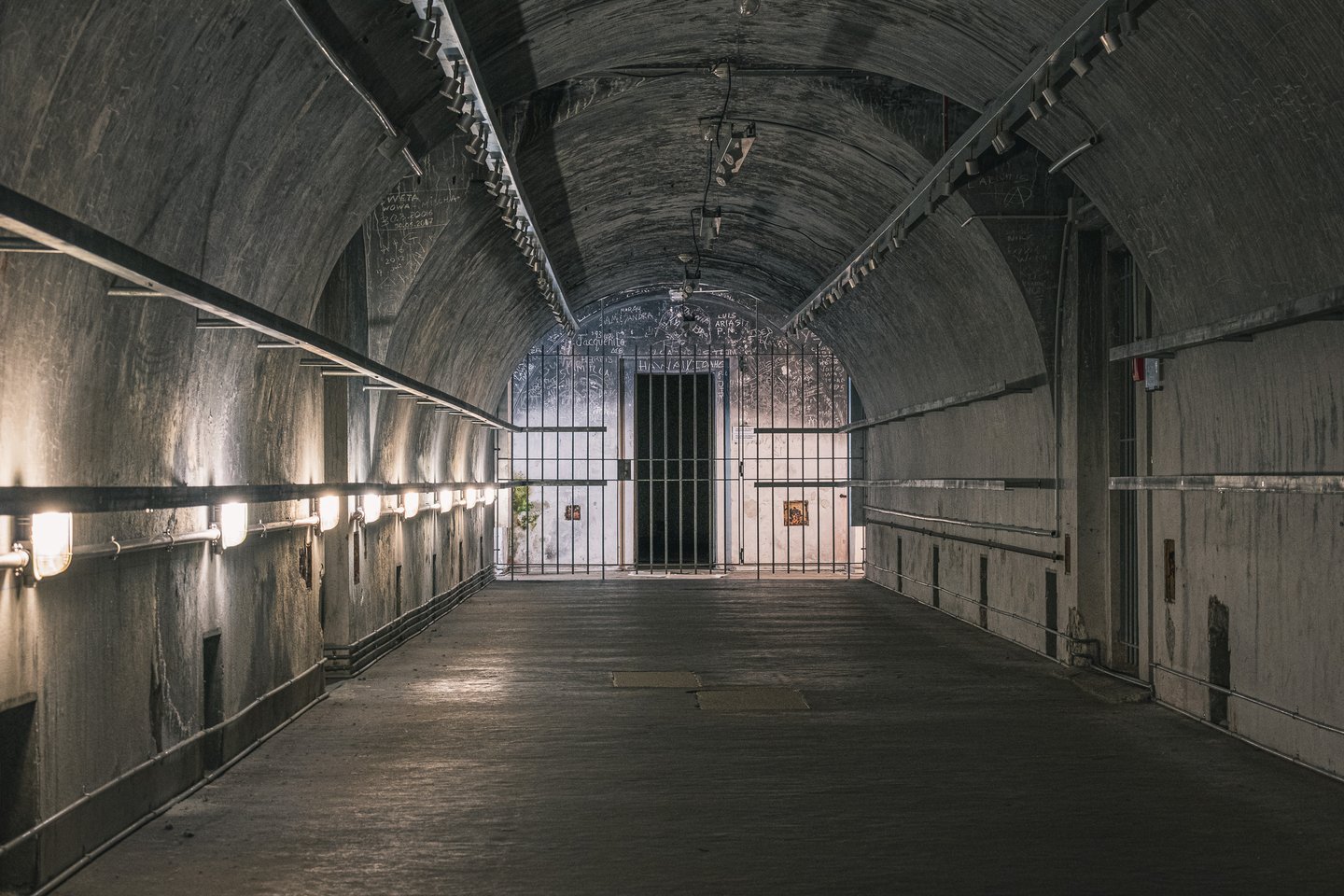 A long barrel vaulted concrete corridor in the Obersalzberg bunker, lit by bulkhead lamps along the left wall and closed at the end by a floor to ceiling steel grille in front of a doorway, the walls around it covered in scratched visitor graffiti.