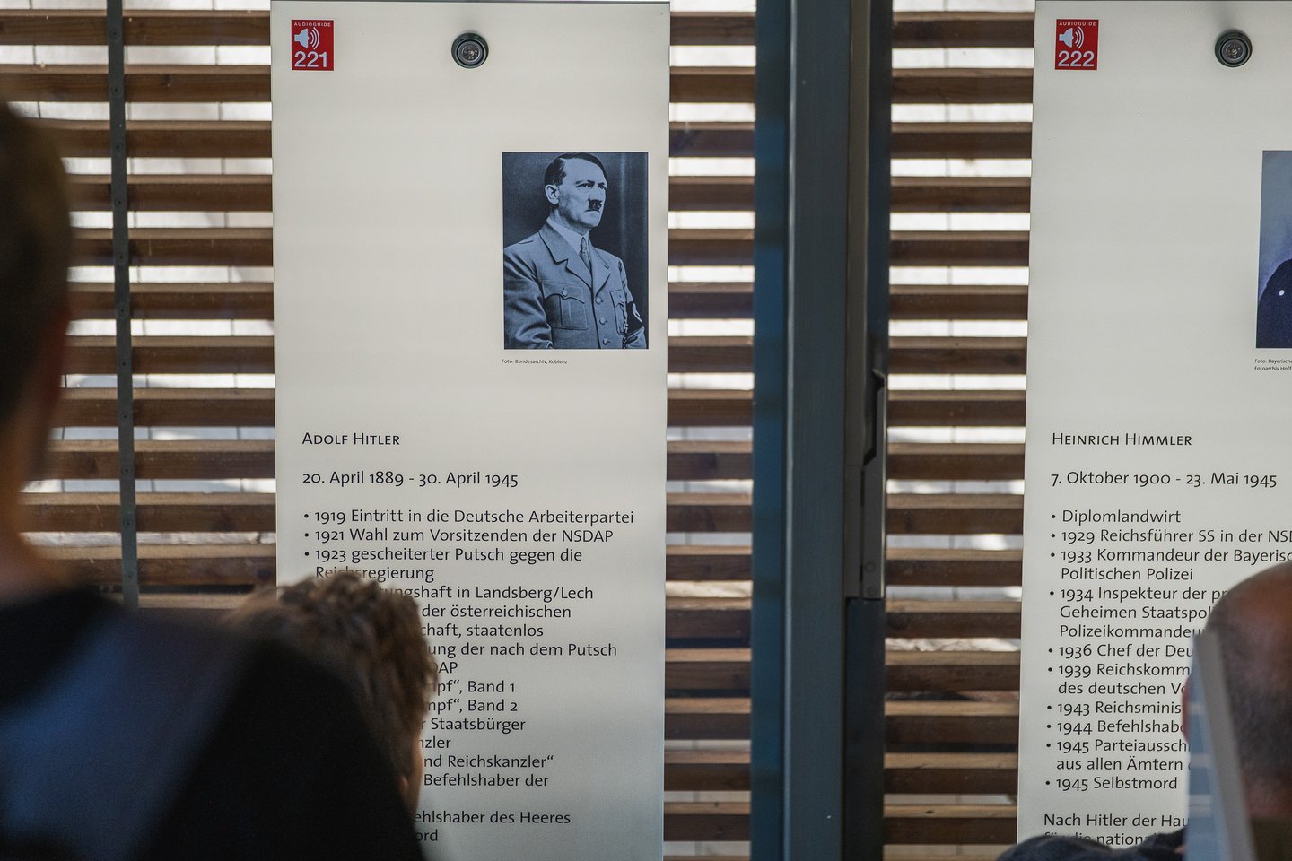 Two tall white exhibition panels inside the Dokumentation Obersalzberg carry biographical timelines of Nazi leaders with red audio guide numbers, seen over the heads of visitors against a slatted wooden wall.