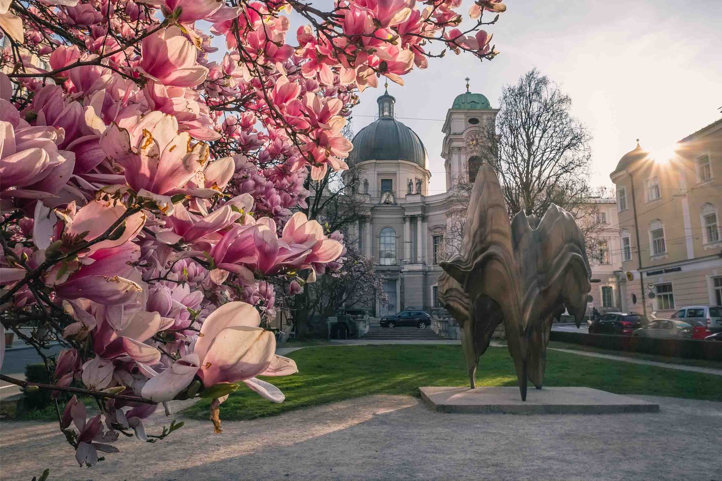 Pink magnolia blossoms and modern bronze sculpture in front of Dreifaltigkeitskirche in Salzburg.