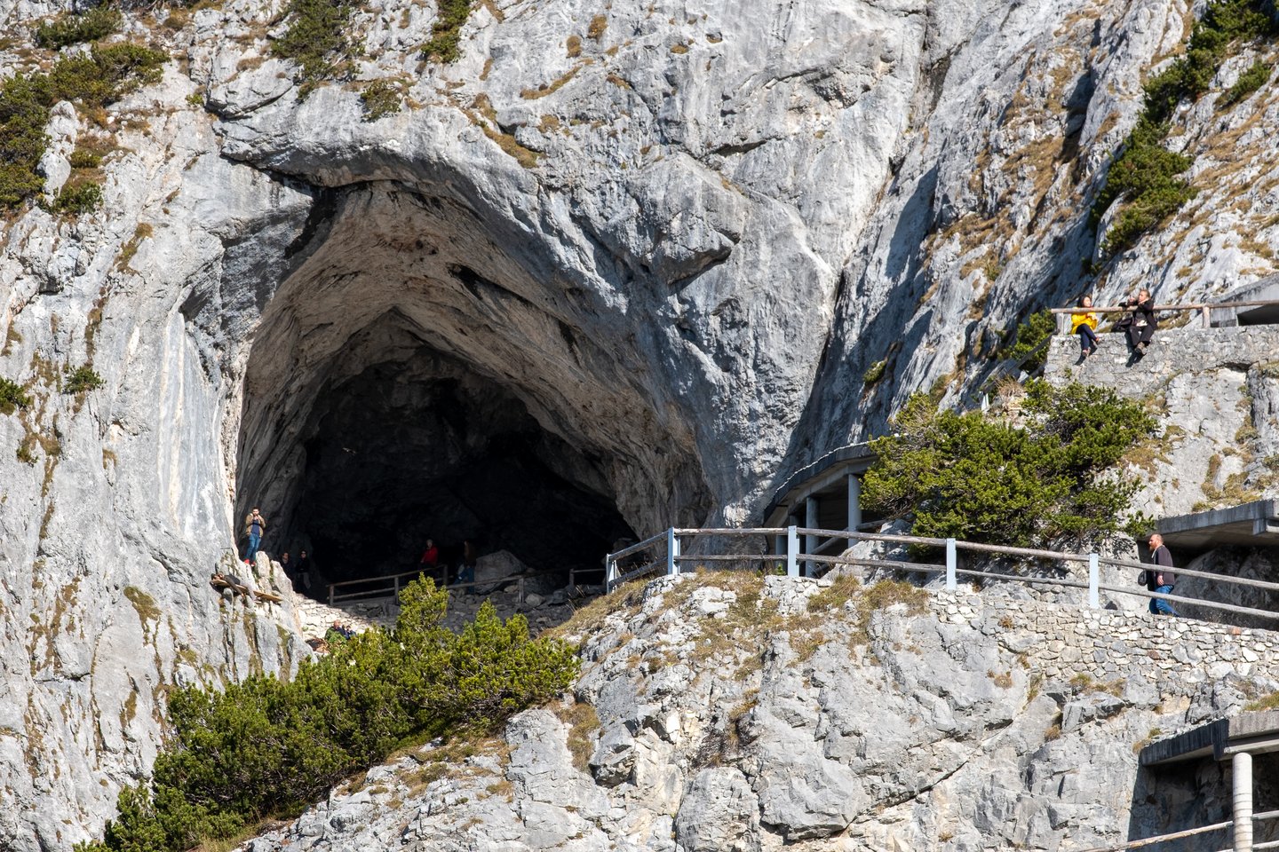 The huge natural rock portal of the Eisriesenwelt ice cave seen from the approach path, with visitors small inside the dark opening.