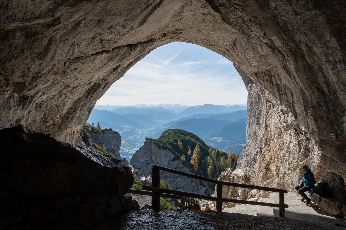 The great arch of the Eisriesenwelt cave mouth frames the Salzach valley and distant alpine ridges, with a lone visitor in blue resting on a bench at the right.
