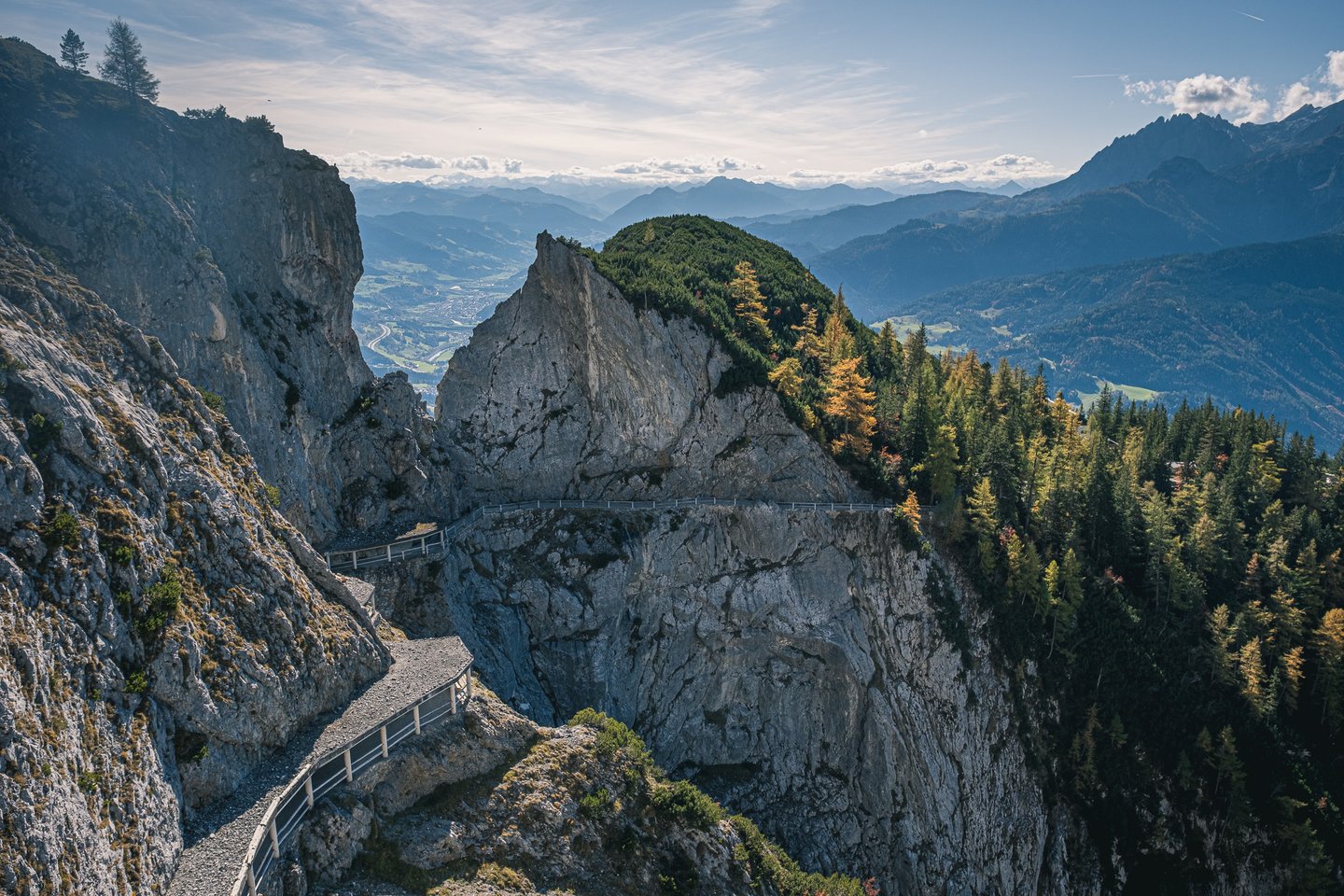 The railed cliff path below the Eisriesenwelt cave entrance winding along a grey limestone face, with larches turning gold and the Salzach valley hazy far below.