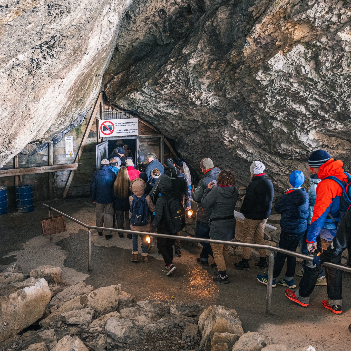 A tour group with lit carbide lamps queues at the wooden inner door of the Eisriesenwelt, under a sign forbidding photography inside the cave.