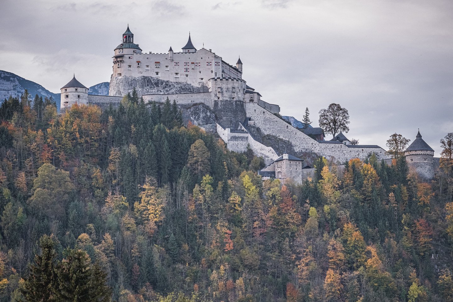 Festung Hohenwerfen sits white on its rock above a hillside of orange and green autumn forest, its keep, clock tower and ring of round bastion towers set against a flat grey sky.