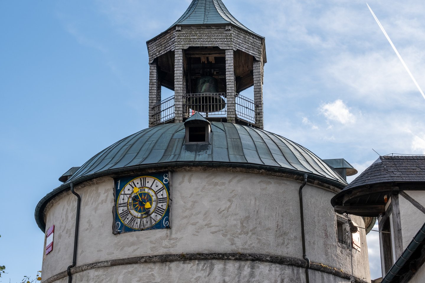 The round tower at Festung Hohenwerfen seen from below against a blue sky, its painted clock face with roman numerals set under a lead dome and an open shingled cage holding the bronze bell.