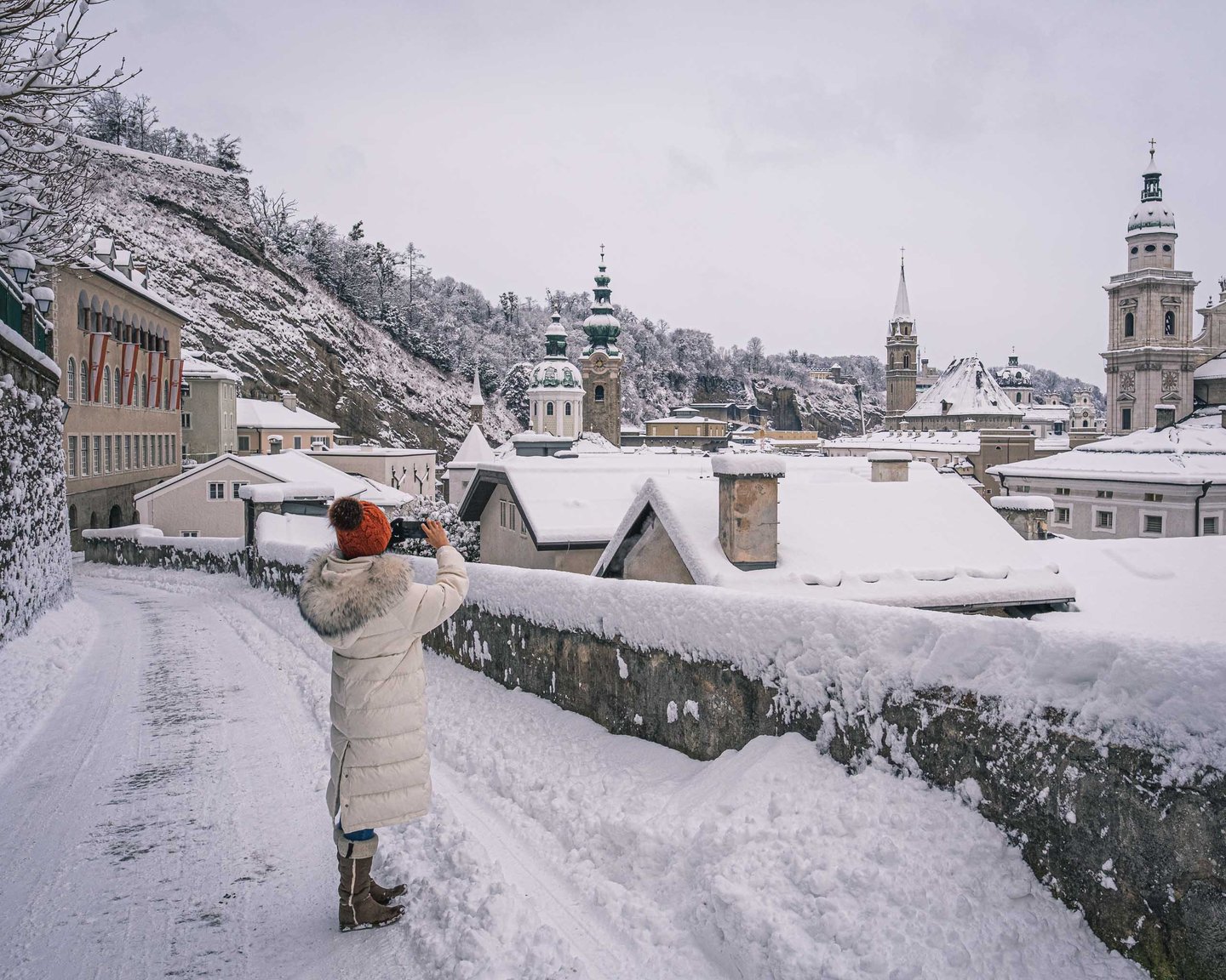 Person in orange hat photographs the snowy Salzburg old town with St Peter and cathedral towers from Festungsgasse.