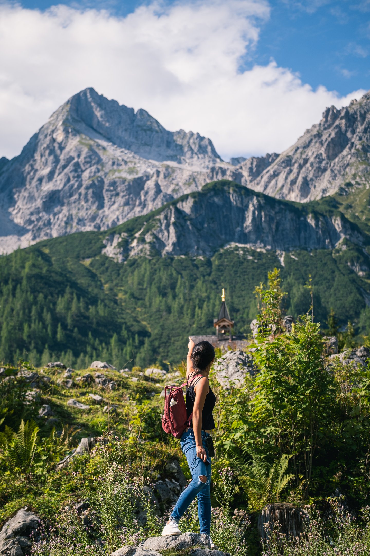 A hiker with a burgundy daypack stands on a rock and points up at a sharp limestone peak above a small shingled chapel tower in Filzmoos.