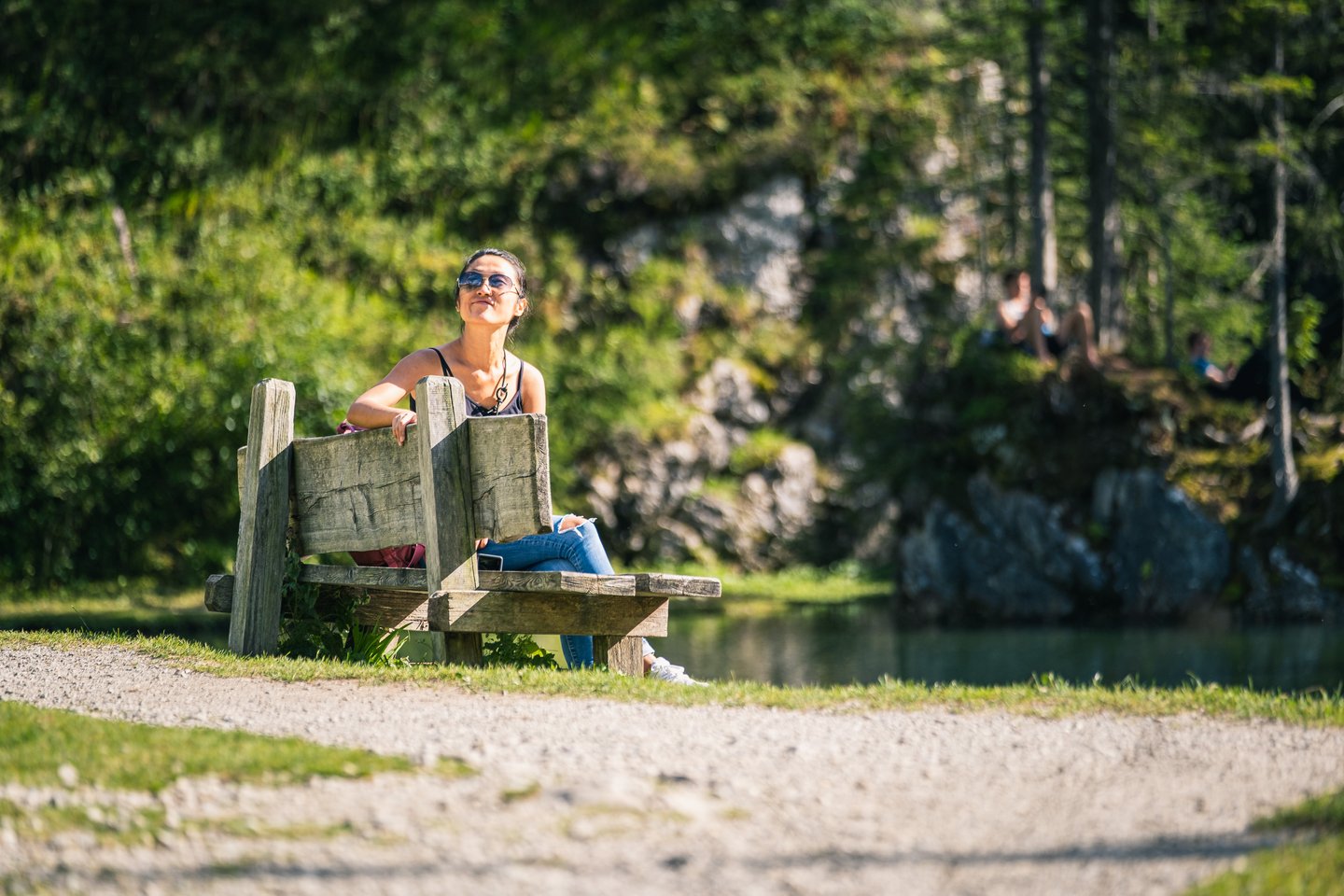 A woman in sunglasses sits back on a rustic timber bench beside a small dark mountain lake in Filzmoos, with a gravel path in the foreground and sunbathers on the rocks opposite.