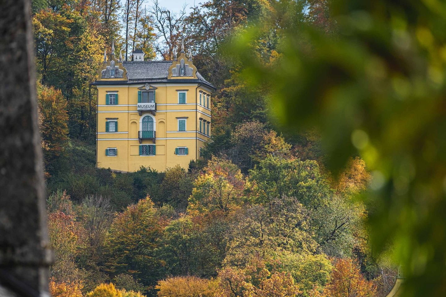 Yellow Baroque Monatsschlossl with museum sign framed by autumn foliage in Hellbrunn park