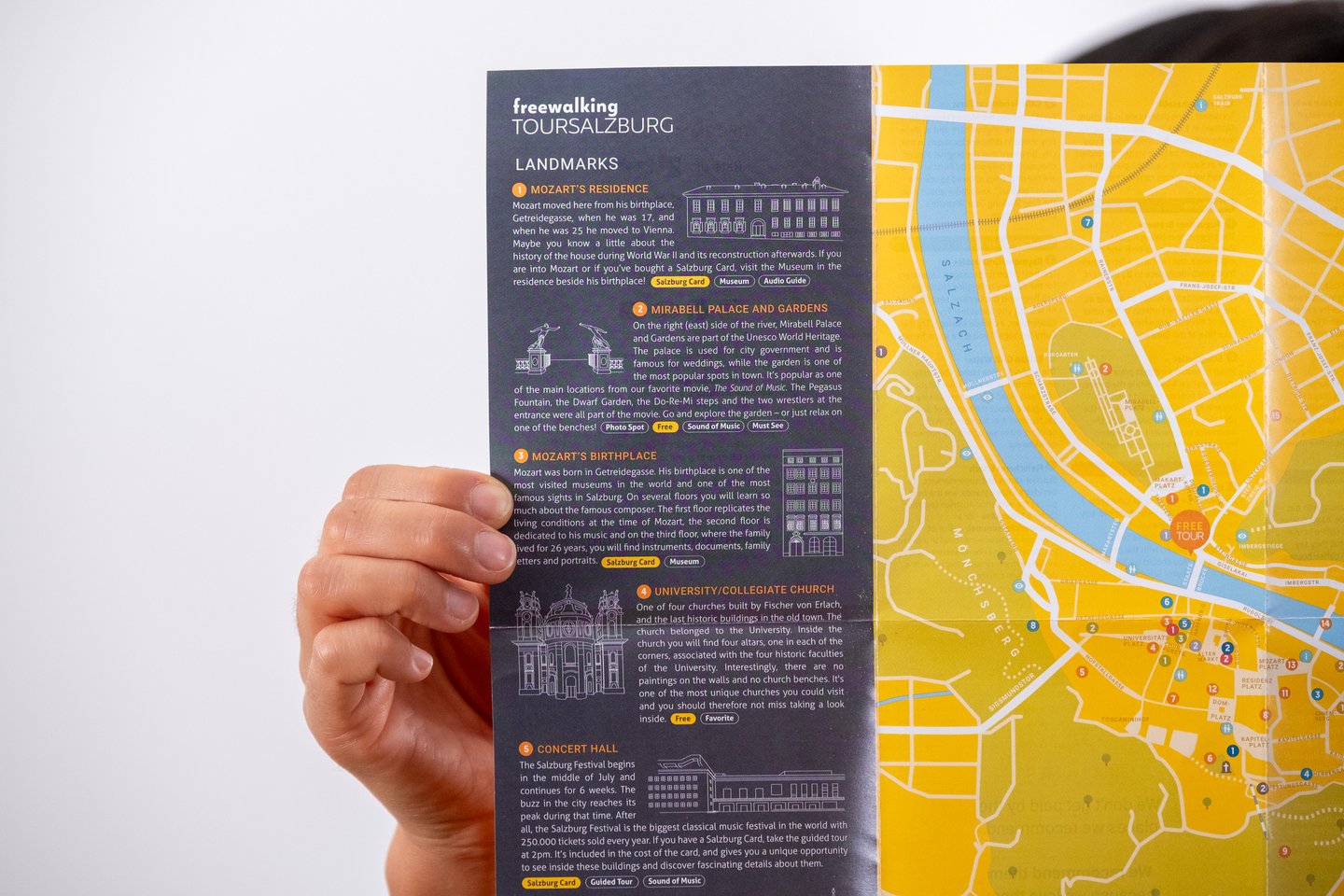 A hand holds the Free Walking Tour Salzburg leaflet open at the landmarks page, with the yellow street map of the old town and the Salzach alongside it.