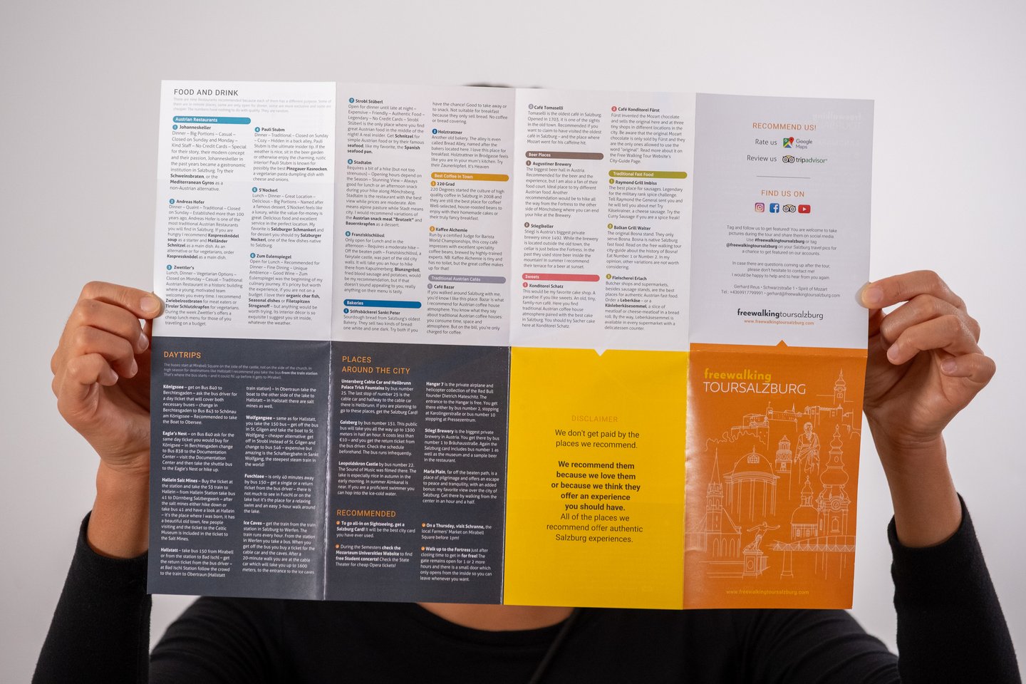 Two hands hold the Free Walking Tour Salzburg city guide open at full spread, showing the food and drink recommendations, the day trips column and the orange cover panel.