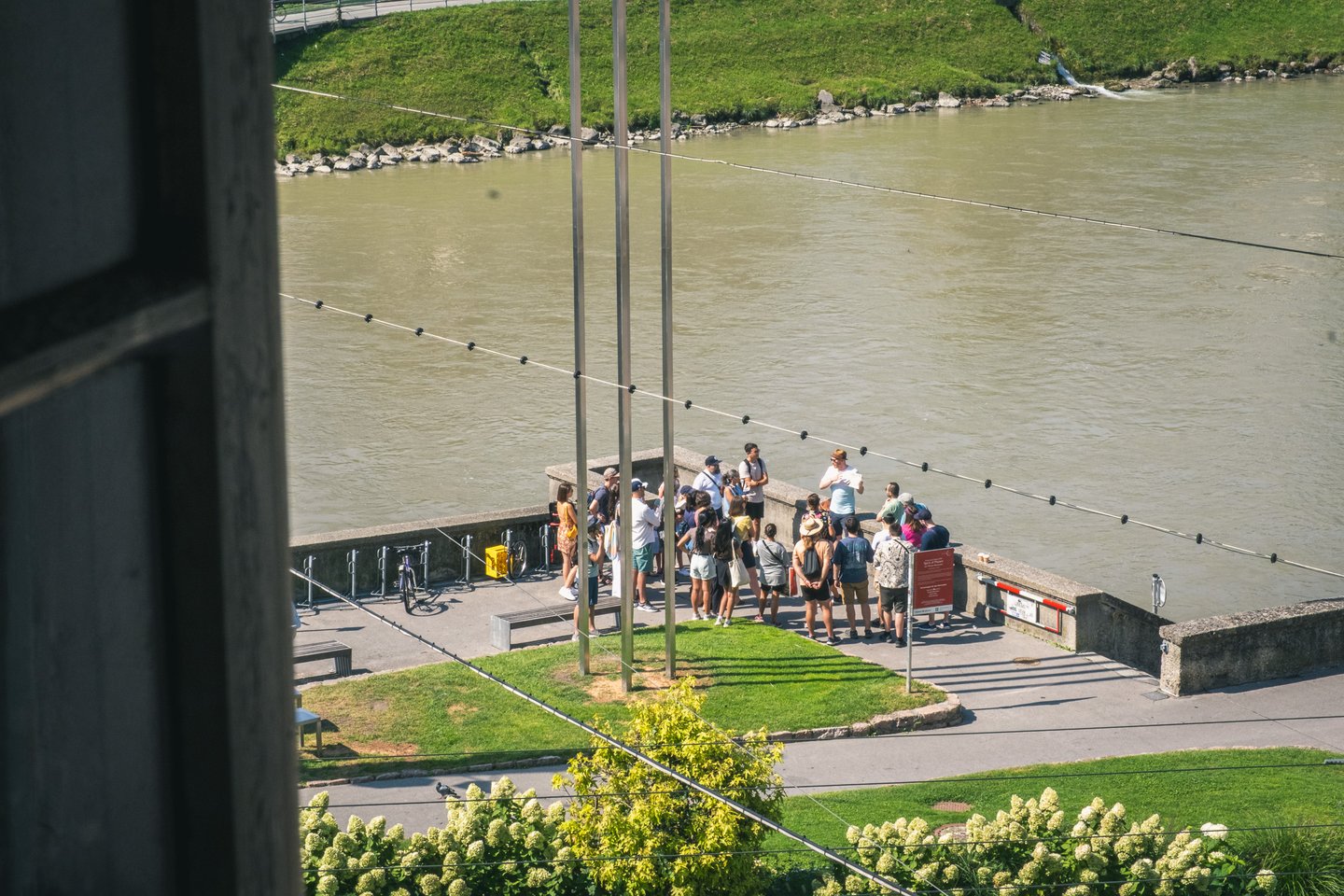 Seen from high above, a summer walking-tour group gathers around a guide in a straw hat on the Salzach embankment beside three tall steel poles.