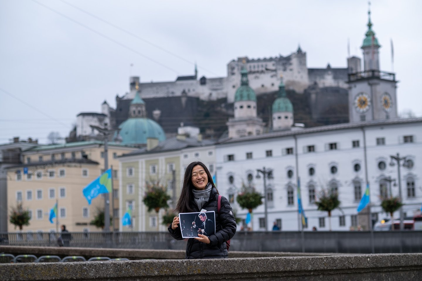 The tour guide laughs as she holds up a printed photograph of conductor Herbert von Karajan mid-beat, with Hohensalzburg Fortress and the cathedral domes behind her.