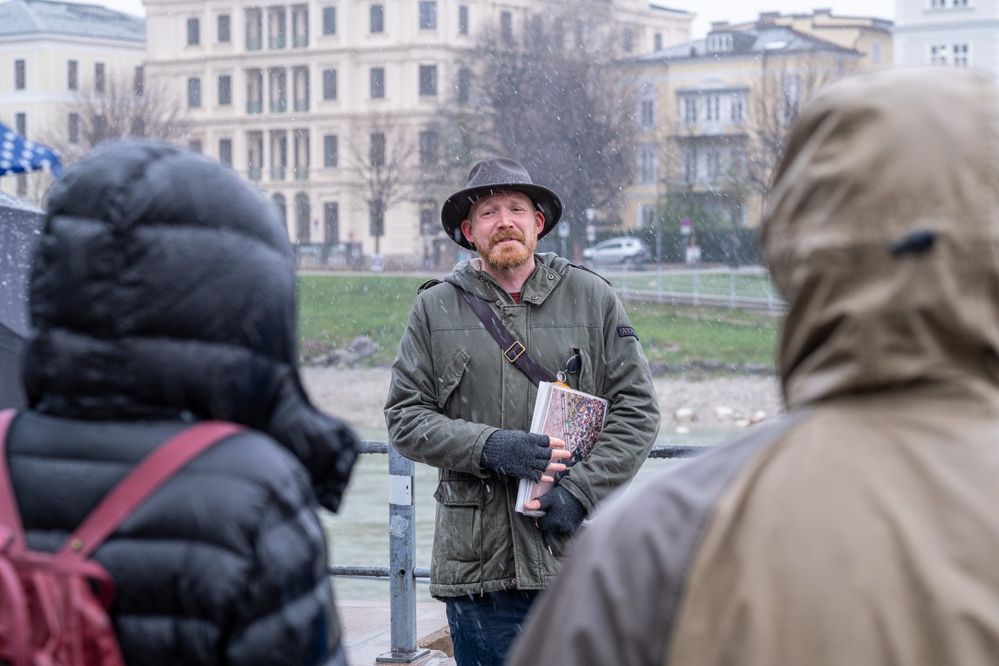 A red-bearded guide in a felt hat and green parka holds his picture book against his chest and speaks to hooded listeners as snow falls beside the Salzach, pale riverside houses opposite.
