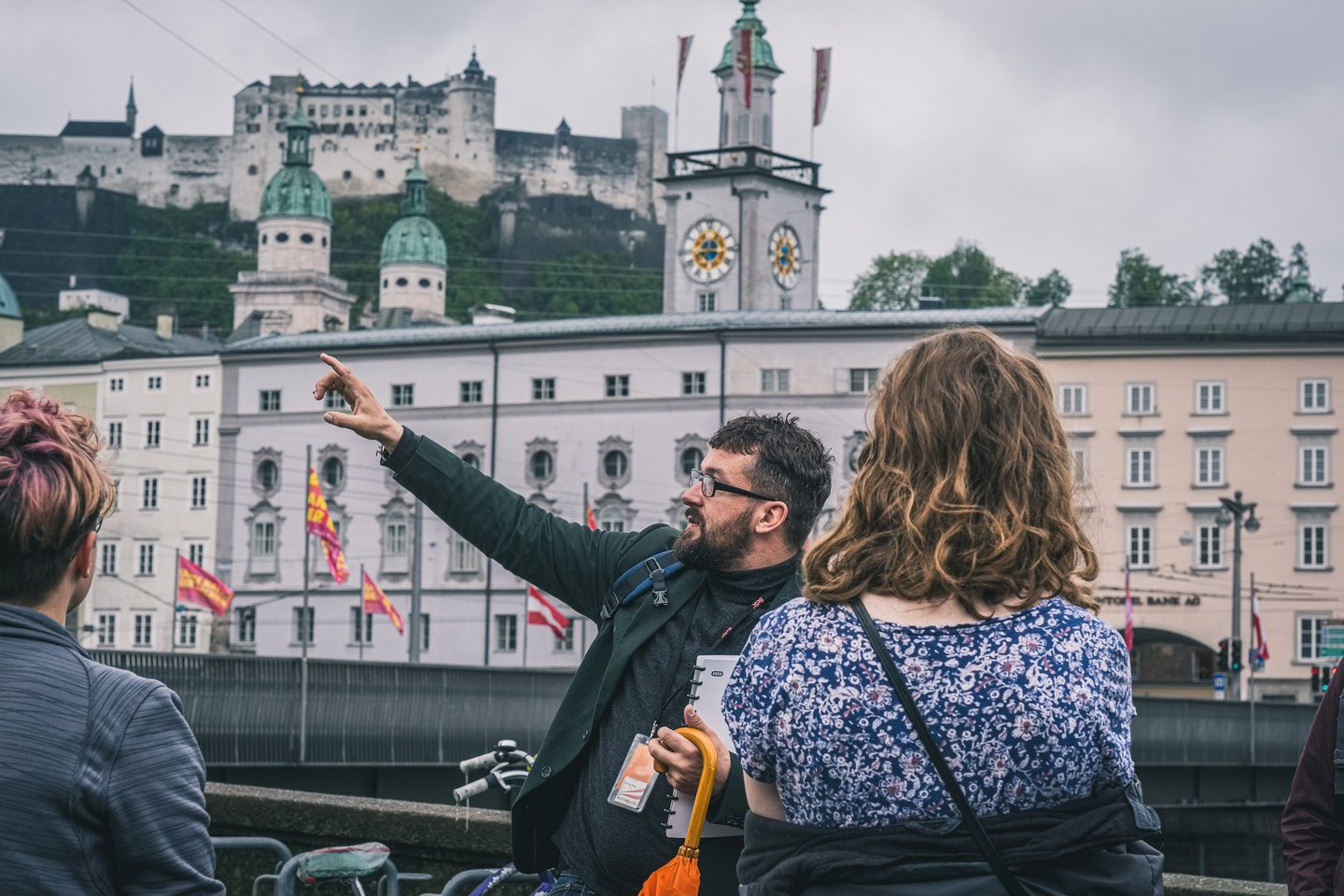 A tour guide with a lanyard and orange umbrella points across the Salzach towards Hohensalzburg Fortress and the town hall clock tower while two guests follow his arm.