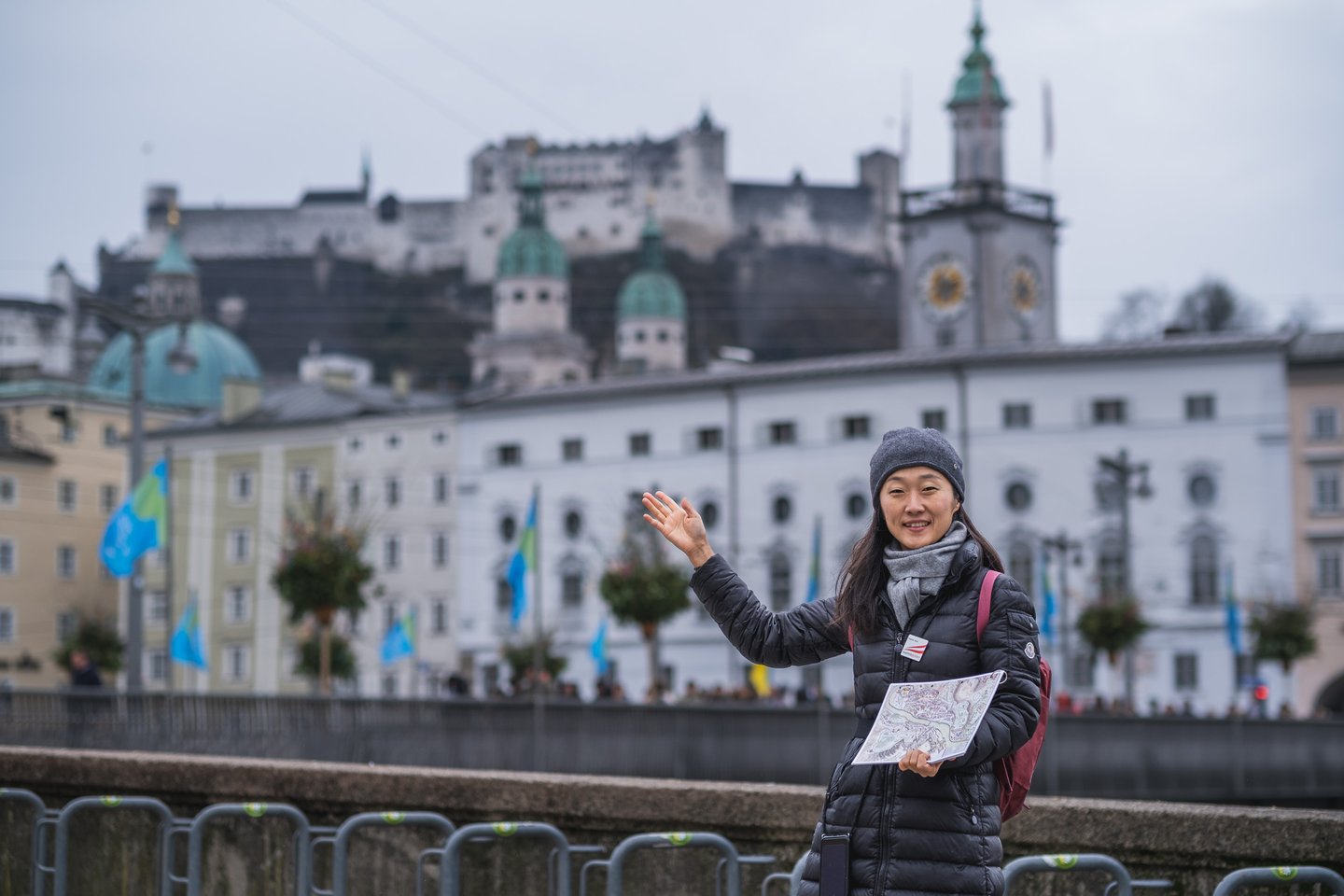 A smiling tour guide in a grey beanie waves while holding the folded Free Walking Tour Salzburg city map, with Hohensalzburg Fortress and the cathedral domes rising behind her.