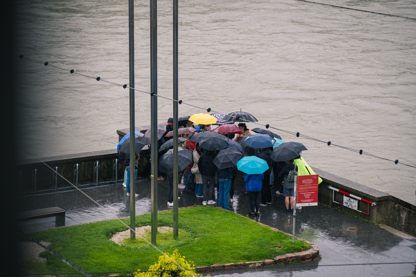 A walking tour group presses together under a cluster of umbrellas in heavy rain on the Salzach embankment, the brown flood water running high behind them.