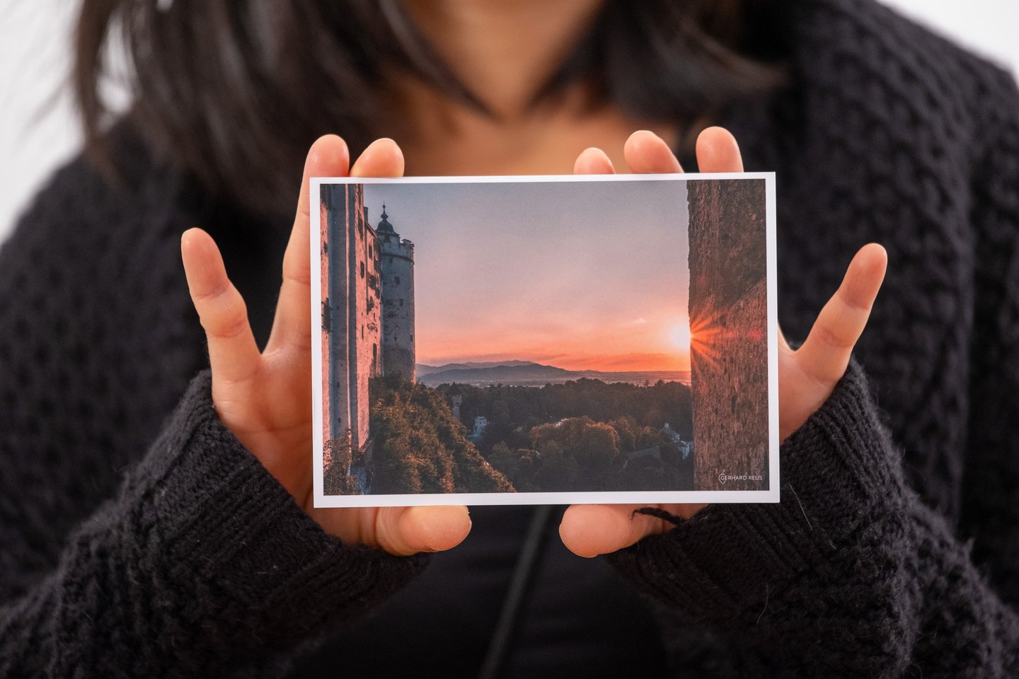 A woman in a dark knitted jumper holds out a postcard showing Hohensalzburg Fortress catching the last orange light of sunset above the trees.