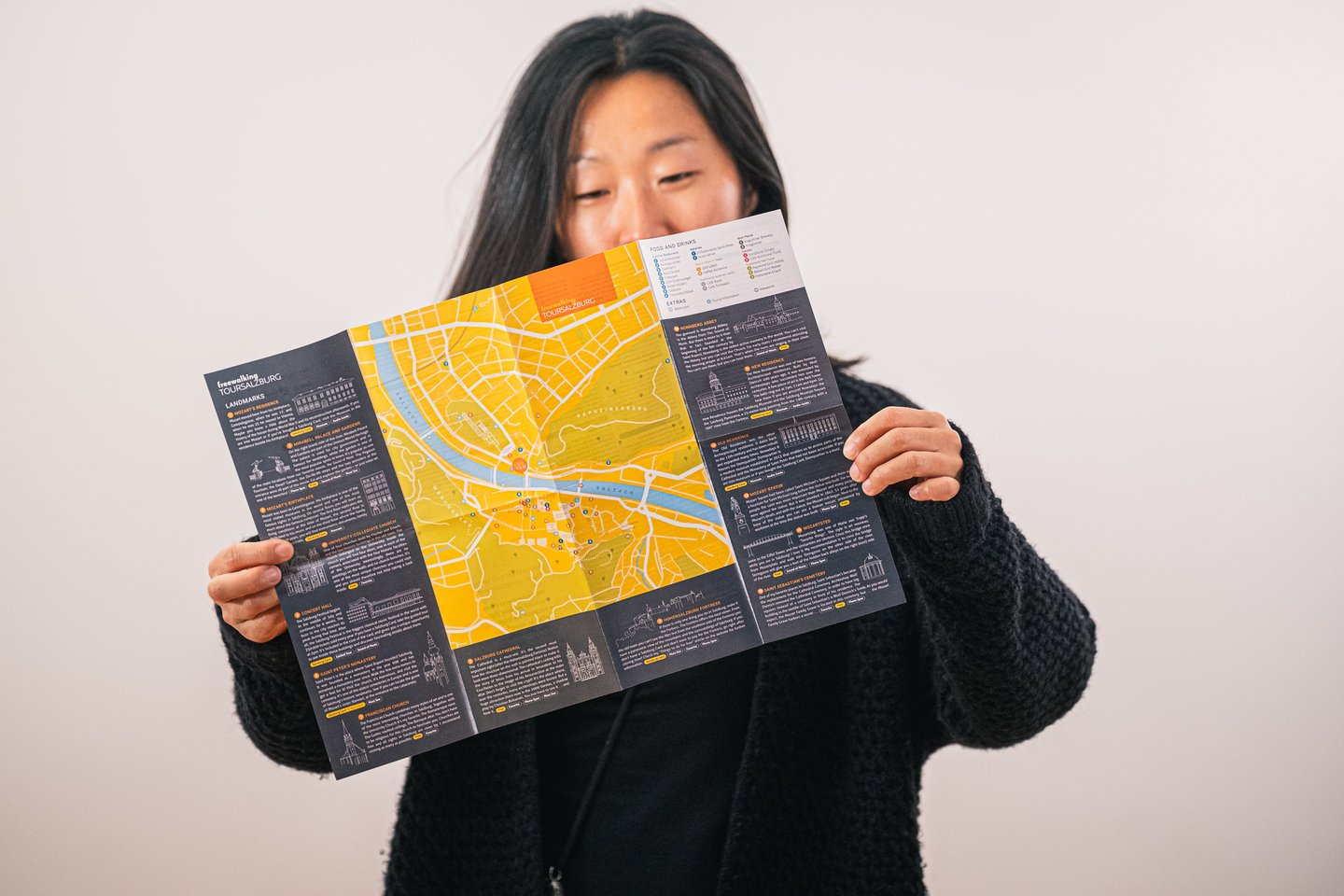 A smiling woman holds open the printed Free Walking Tour Salzburg city map, its yellow street plan of the old town flanked by illustrated landmark notes.