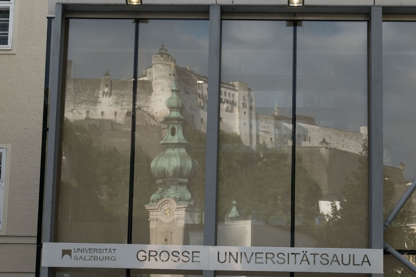 Fortress and Franciscan Church tower reflected in Grosse Universitaetsaula glass facade in Salzburg