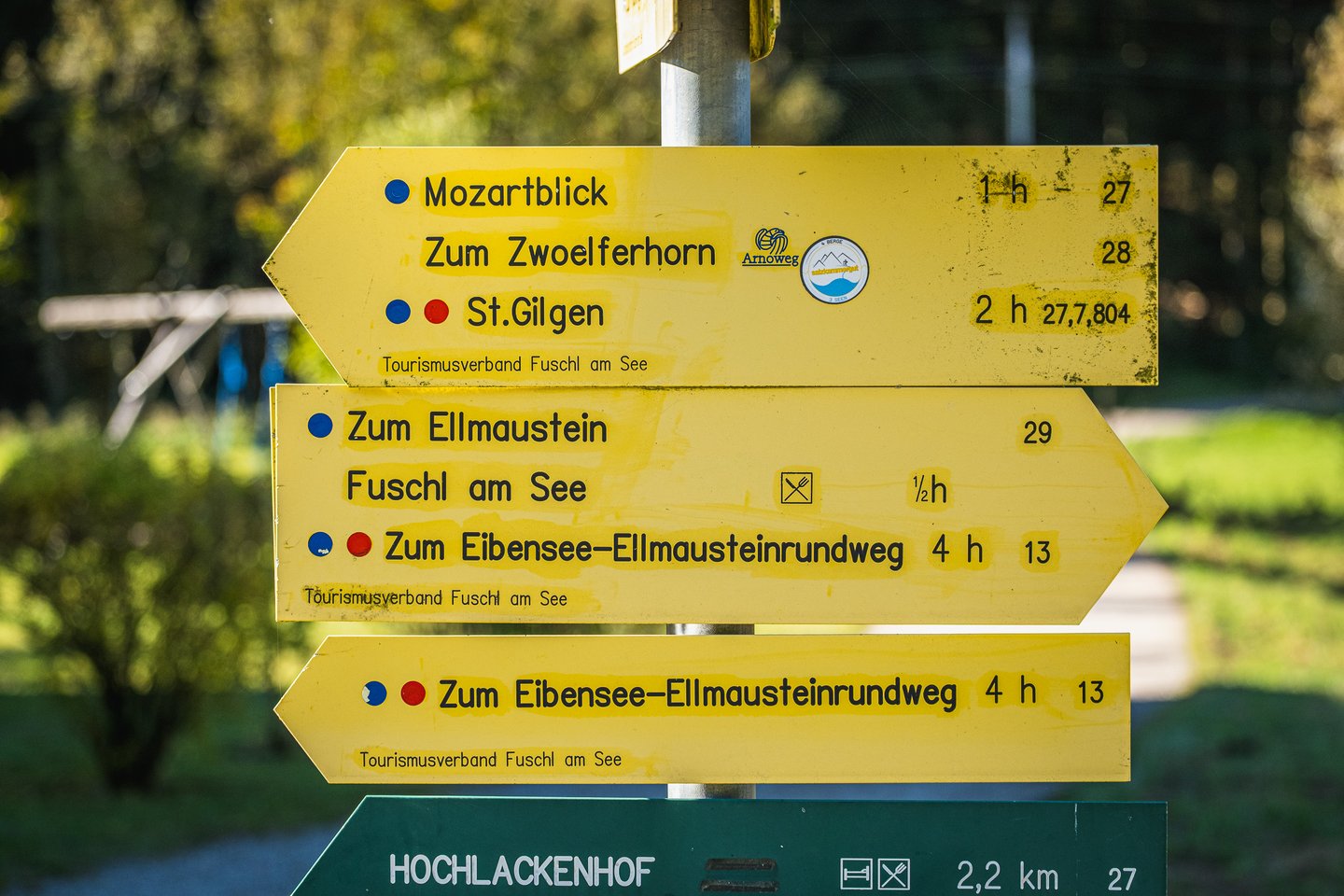 A yellow Fuschl am See signpost lists Mozartblick, the Zwoelferhorn, St. Gilgen and the Ellmaustein circuit, above a green sign for the Hochlackenhof.
