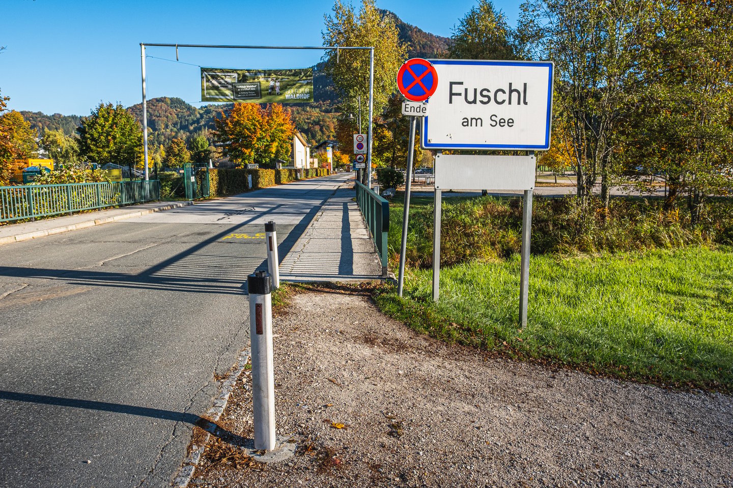The blue-bordered Fuschl am See village sign beside the road into the village on a bright autumn morning, with a golf club banner strung across the street ahead.