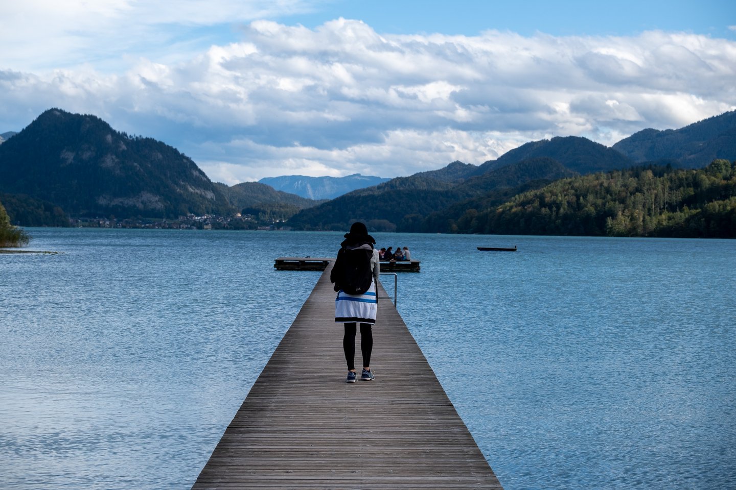 A woman with a backpack walks down the centre of a long wooden jetty on Fuschlsee toward a group sitting at its far end, mountains rising on both sides of the lake.