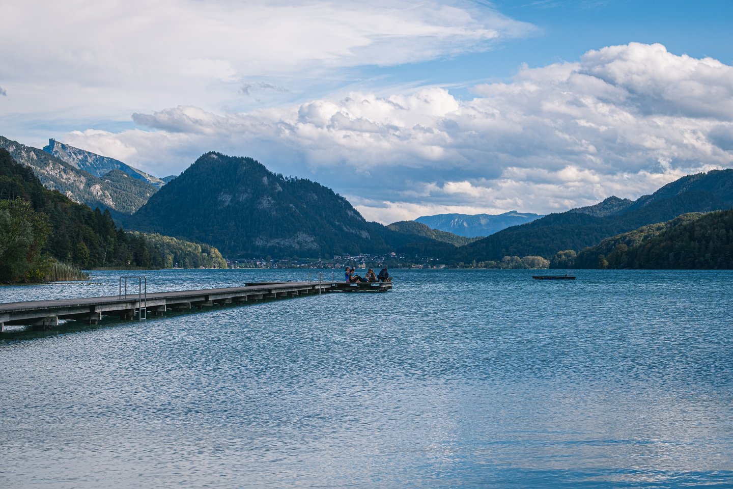 A long wooden jetty reaches far out across Fuschlsee toward the distant village of Fuschl am See, under a wide sky of broken cloud with the Schafberg ridge on the horizon.