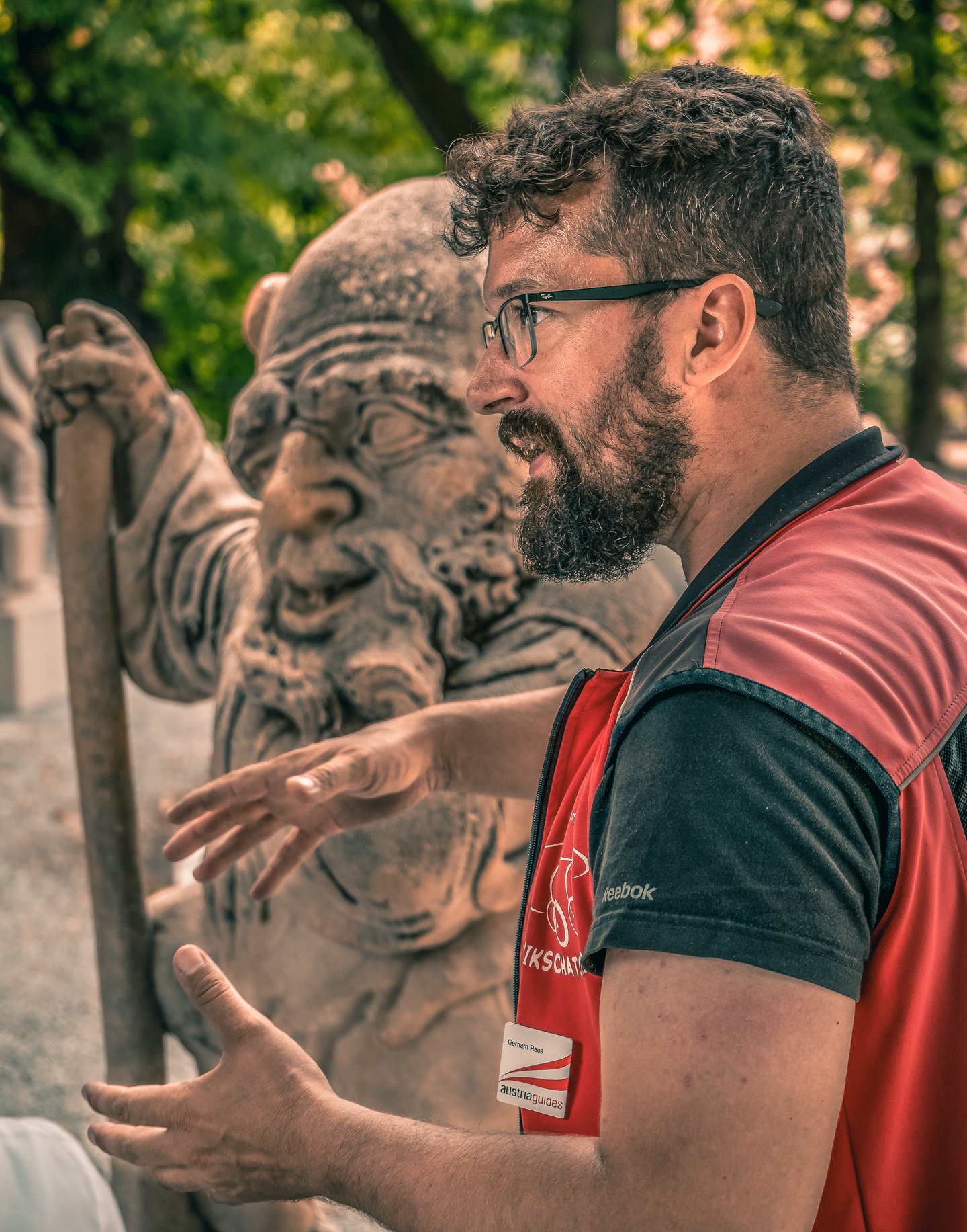 Gerhard Reus talks with both hands open in front of a weathered marble dwarf leaning on a staff in the Zwerglgarten, his austriaguides badge pinned to a red Rikschatours vest.