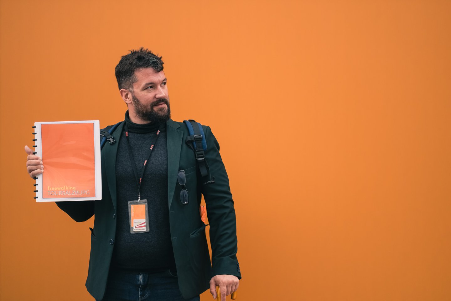 Gerhard Reus, in a dark green blazer with an Austria Guides badge on a lanyard, holds up his orange ring-bound Free Walking Tour Salzburg folder against a plain orange background.