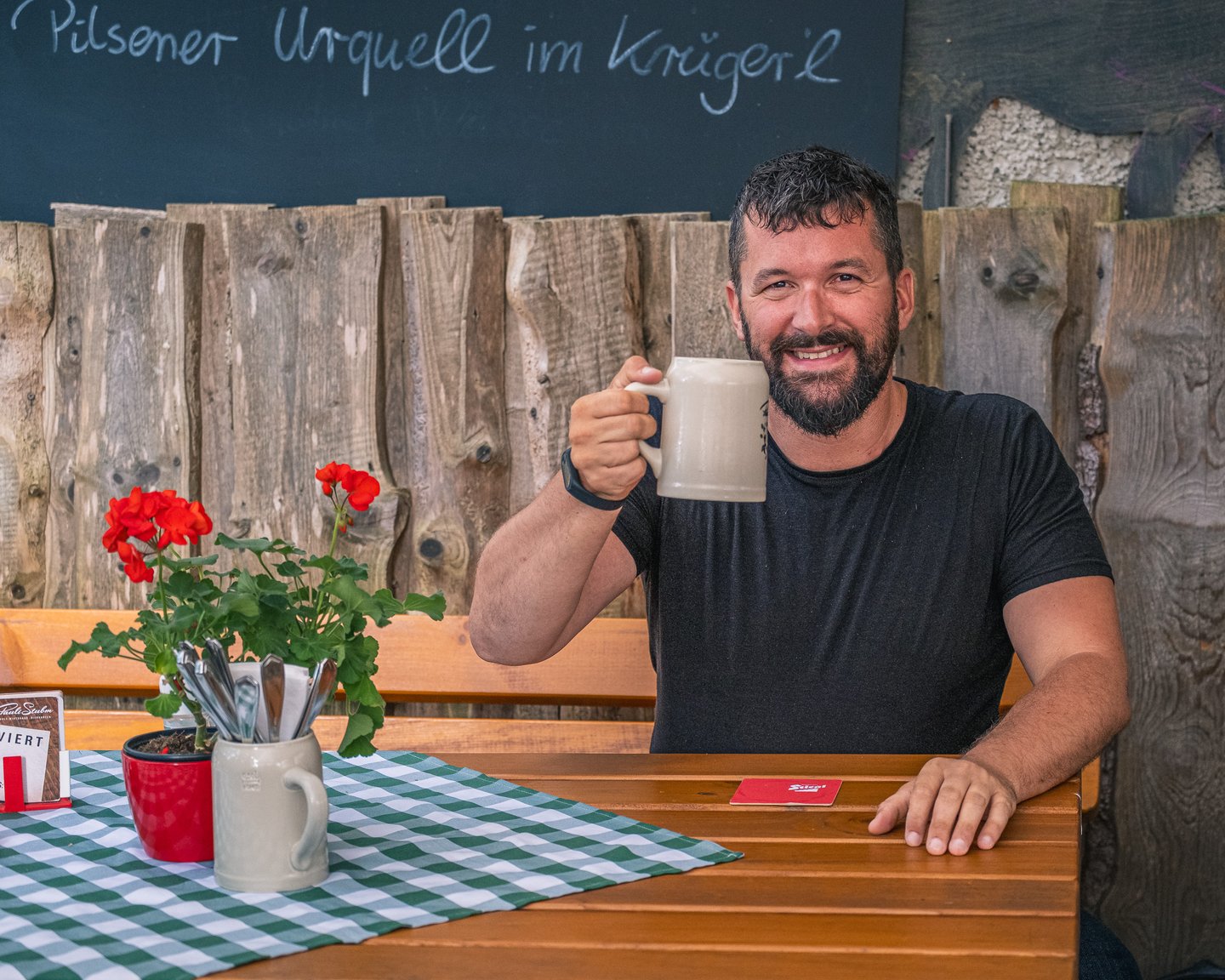 Gerhard Reus, bearded and in a black t-shirt, sits at a beer garden table at Pauli Stubm and raises a stoneware stein towards the camera, with a chalkboard and red geraniums beside him.
