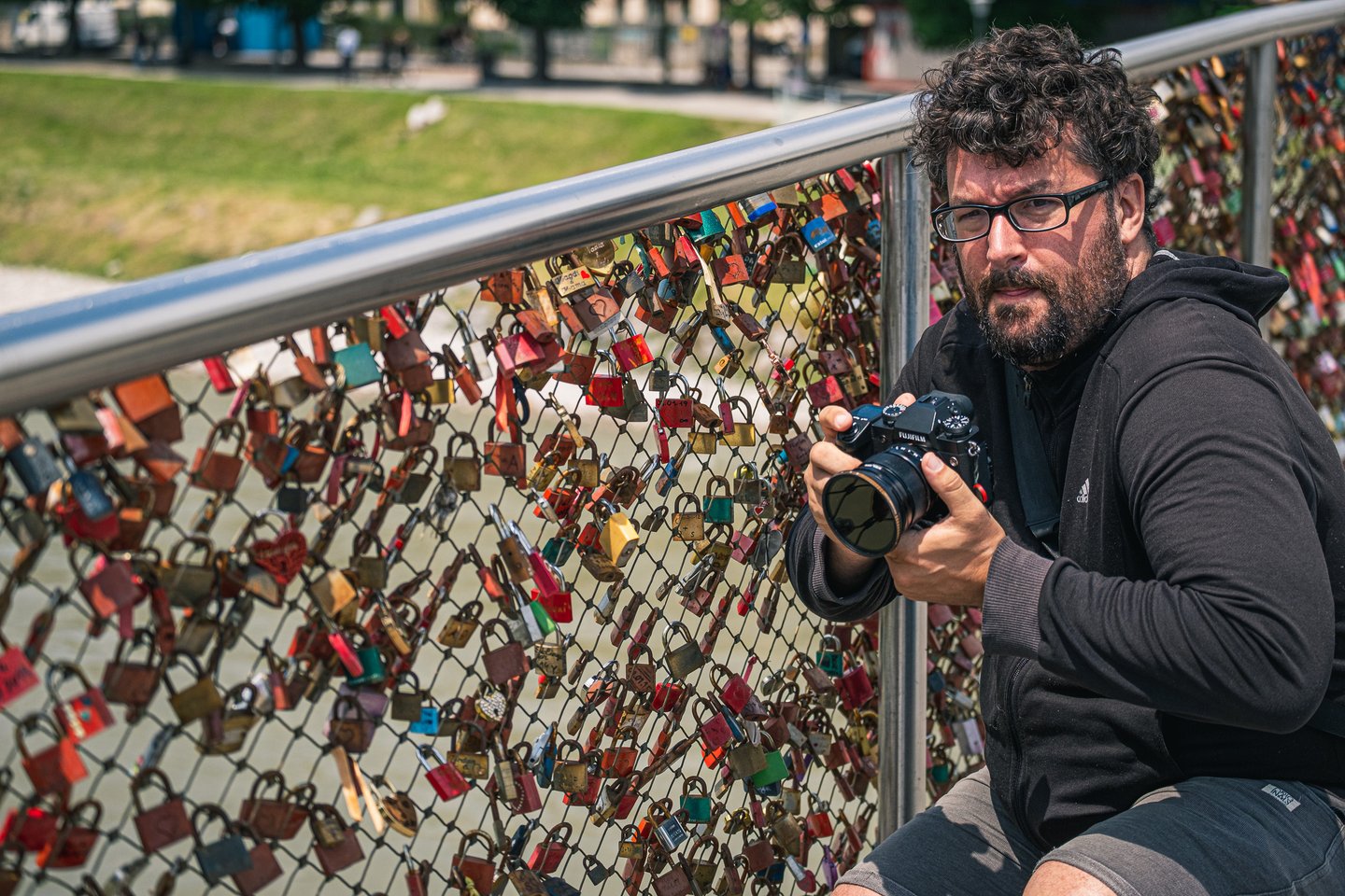 Gerhard Reus, in a black hoodie and glasses, crouches at the love lock railing of the Marko-Feingold-Steg holding a Fujifilm camera and looking towards the viewer.