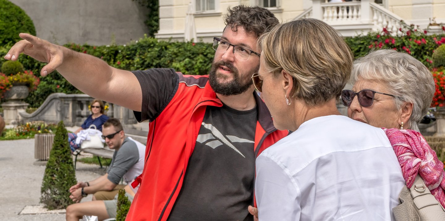 Gerhard Reus, in a red Rikschatours guide vest, stretches out an arm to point something out while two women listen, roses and clipped hedges behind him in the Mirabell Gardens.