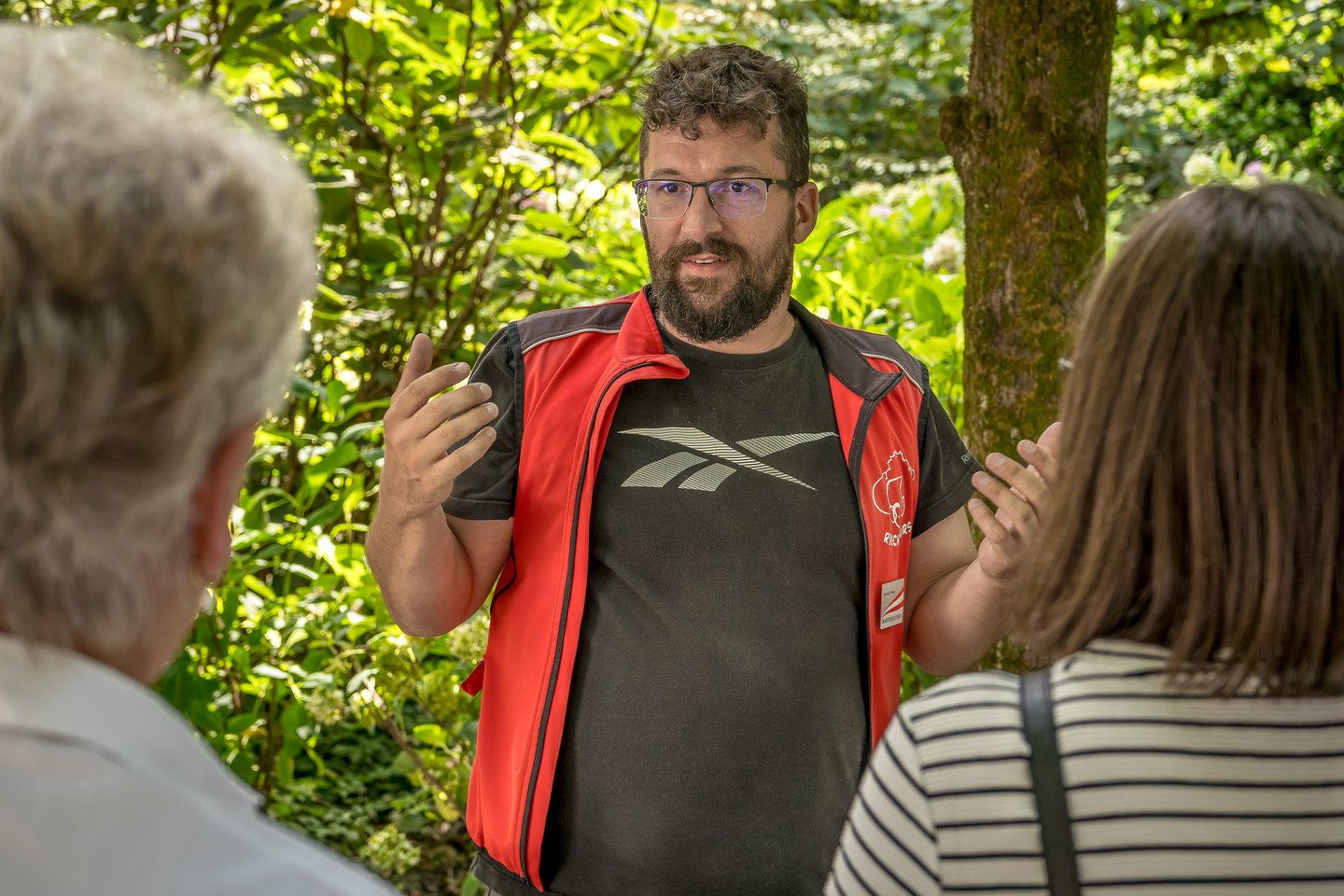 Gerhard Reus speaks with both hands raised, framed head-on between the blurred shoulders of two listening guests, sunlit shrubs and a mossy trunk behind him.