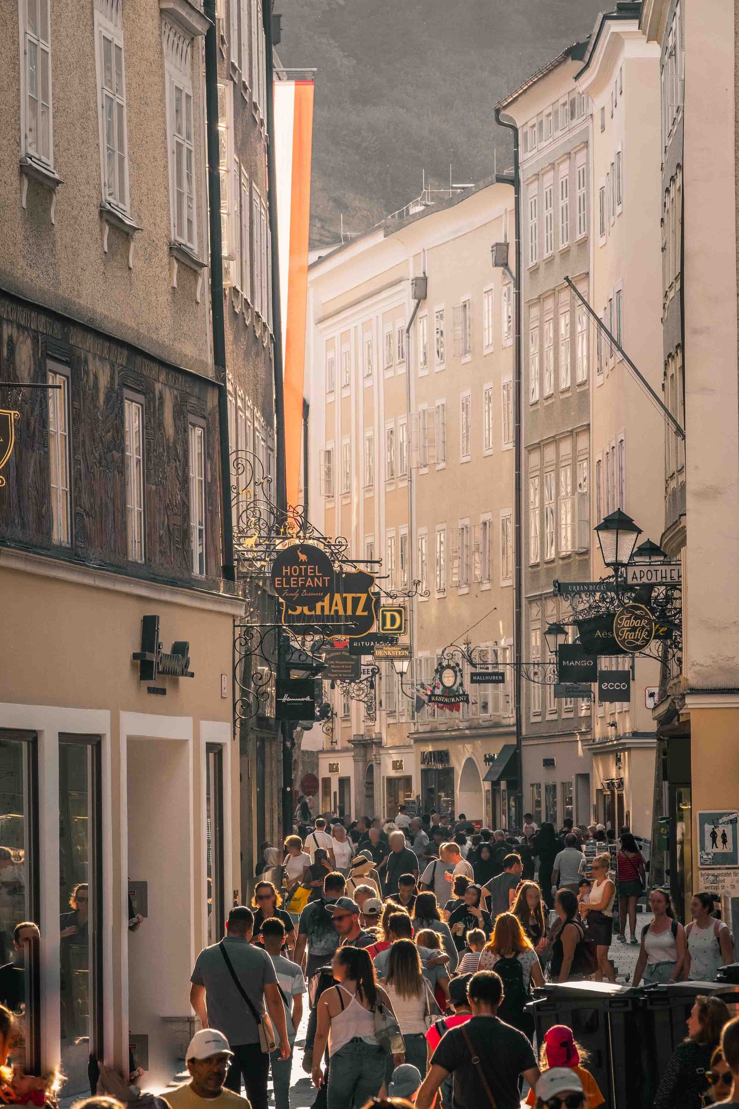 Crowds fill Getreidegasse with the wrought-iron Hotel Elefant and Schatz Konditorei signs overhead in Salzburg old town.