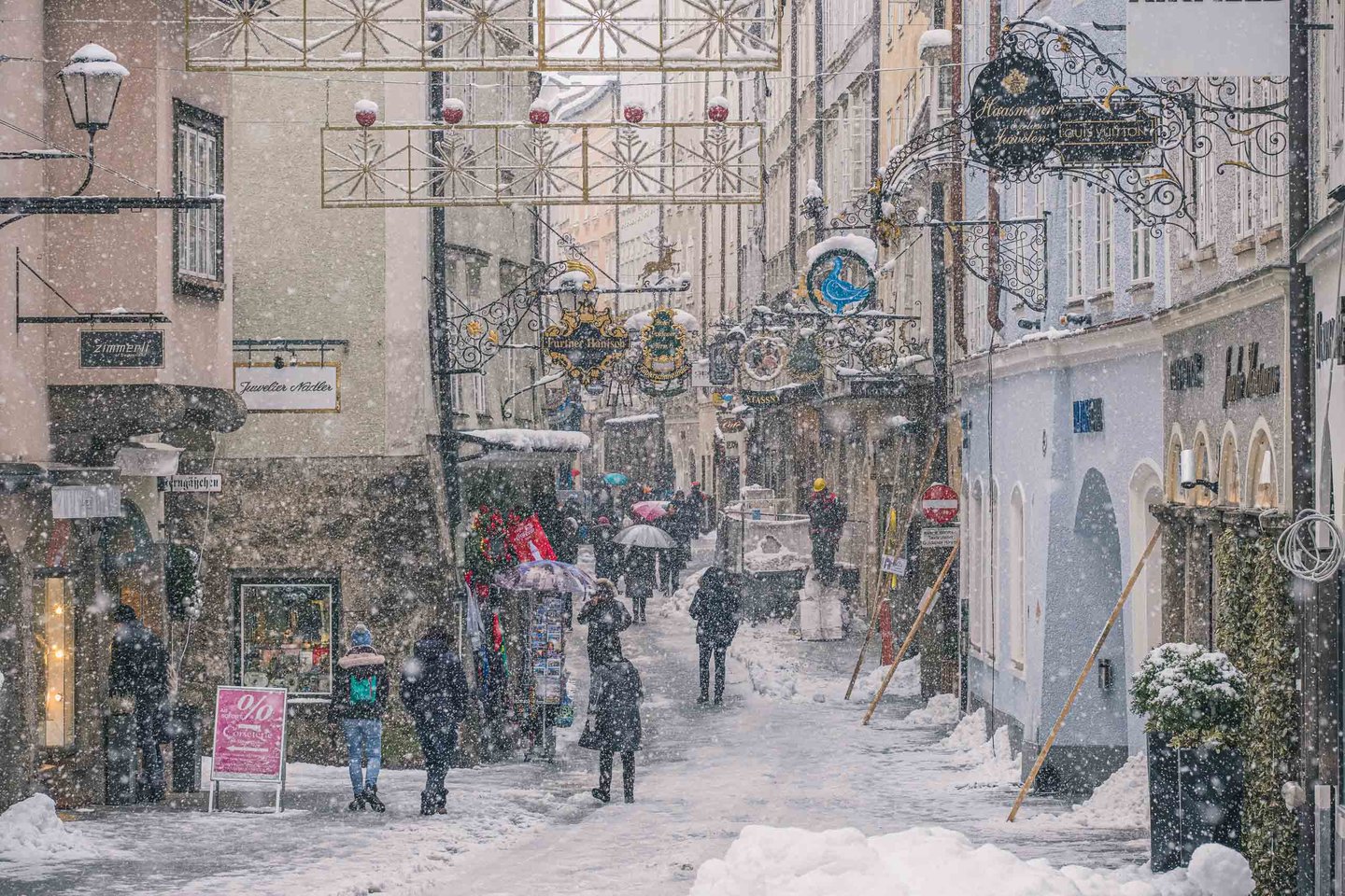 Snow falling on Getreidegasse with wrought iron guild signs and Christmas decorations in Salzburg old town.