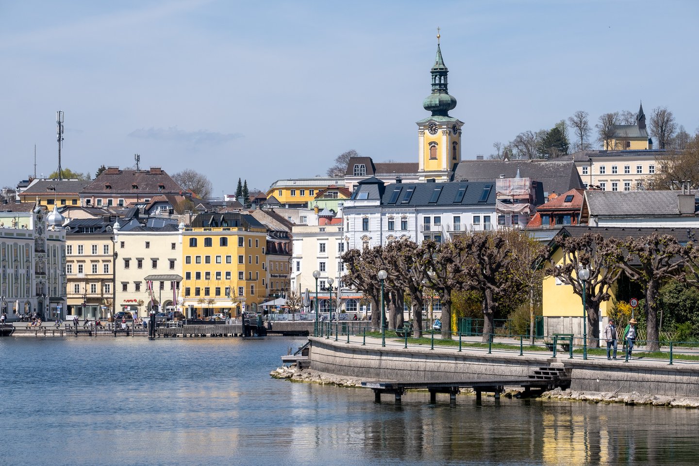 The Gmunden lakefront in spring, with the pastel Rathausplatz houses and the yellow-towered parish church on the left and the Esplanade promenade curving along the right behind pollarded trees, all seen from the Traunsee.