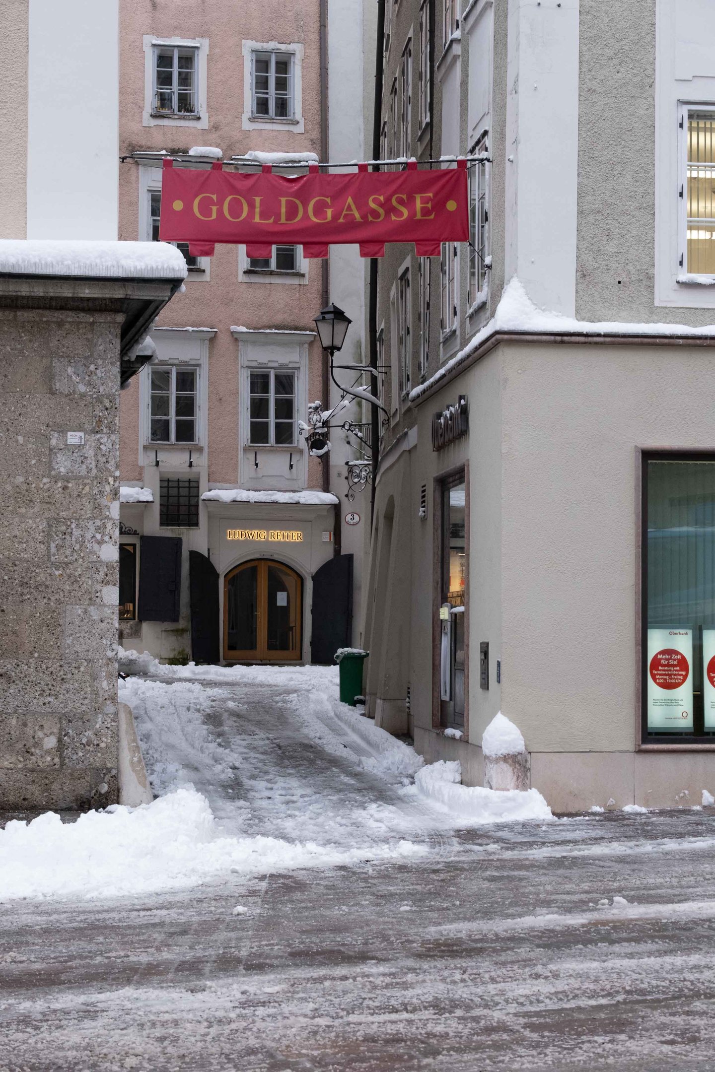Red Goldgasse banner hangs over a snowy narrow lane in Salzburg old town in winter.