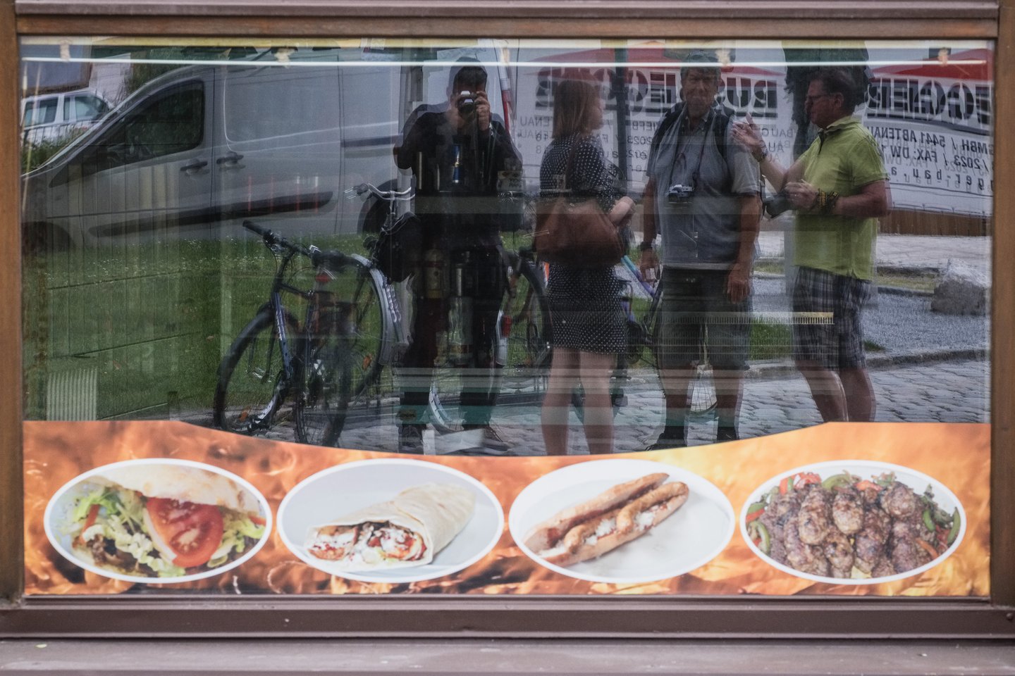 The photo menu strip in a kebab shop window in Hallein, the glass reflecting parked bicycles, passers-by on the cobbles and the photographer raising his camera.