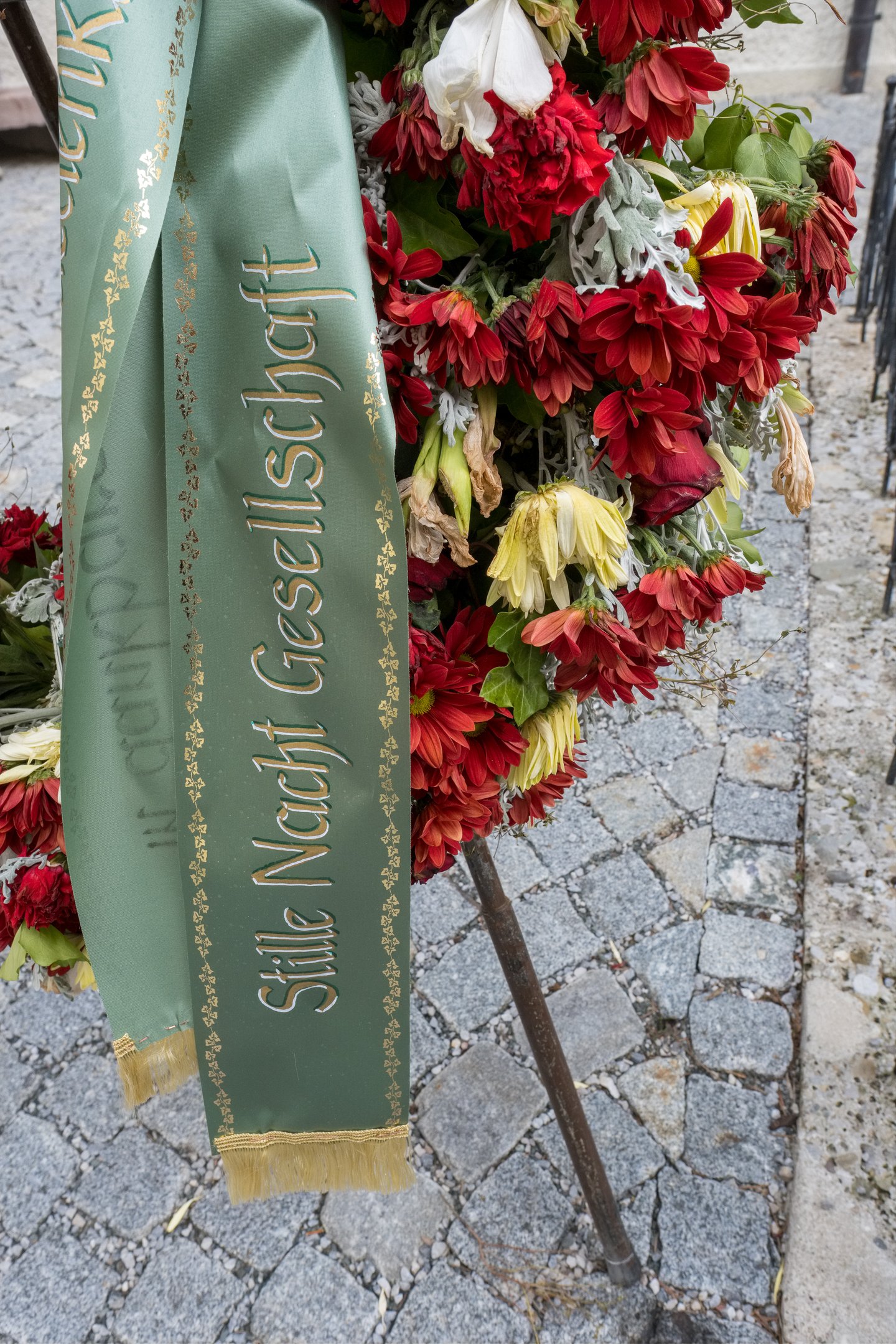 A memorial wreath of red and yellow flowers with a green sash lettered Stille Nacht Gesellschaft in gold, standing on cobblestones in Hallein.