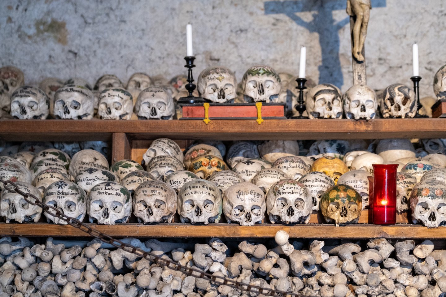 A wide view of the charnel house wall showing three tiers of painted skulls beneath a wooden crucifix and the blue cross painted on the whitewash behind it, with the lowest register filled by packed bones.