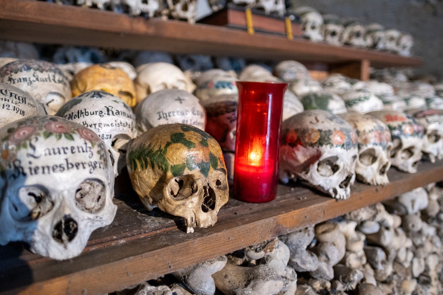 A red glass votive lamp burns at shelf level among the painted skulls, its light falling on a skull crowned with green and yellow leaves and on one lettered Laurentz Rienesberger.