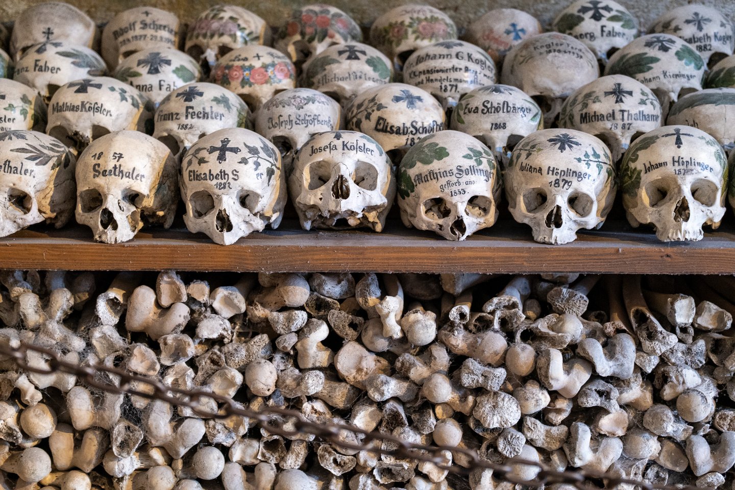 A long row of skulls painted with names, dates, crosses and leafy wreaths rests on a wooden shelf, with the lower half of the frame taken up by a tightly packed wall of thigh bones.